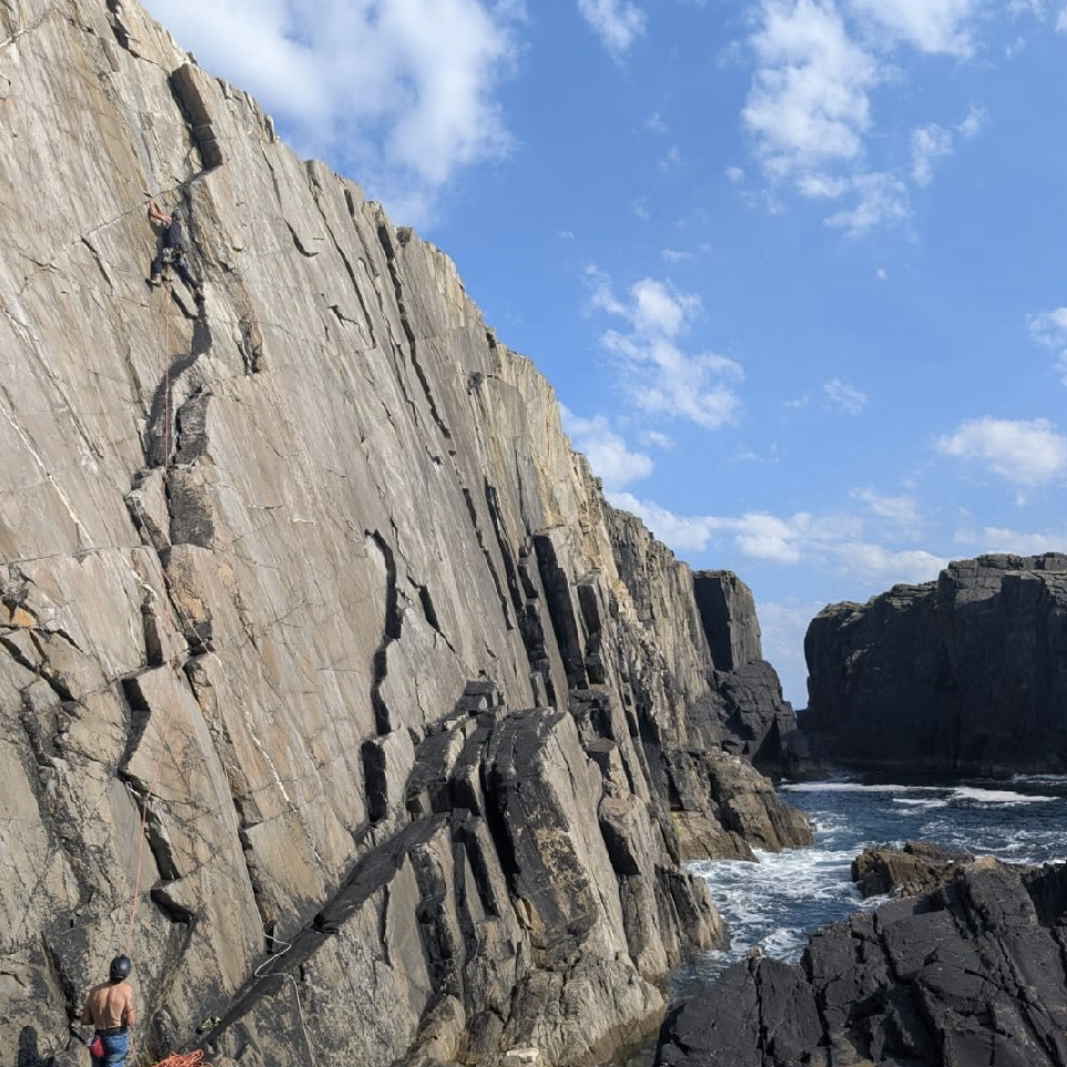 A person climbing a steep rocky cliff beside the ocean, with another person at the bottom preparing to climb, under a partly cloudy sky.