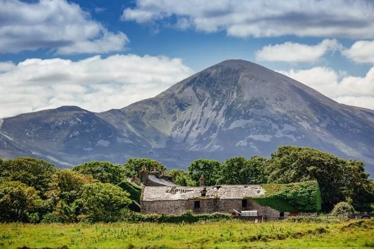 A rural landscape with a stone building surrounded by green trees and grass, with a large mountain in the background under a partly cloudy sky.