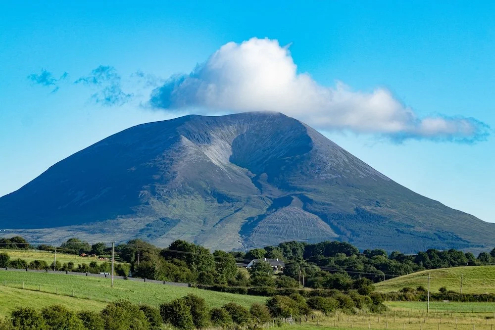 A large mountain with a cloud at its summit, green fields, trees, and power lines in the foreground under a clear blue sky.