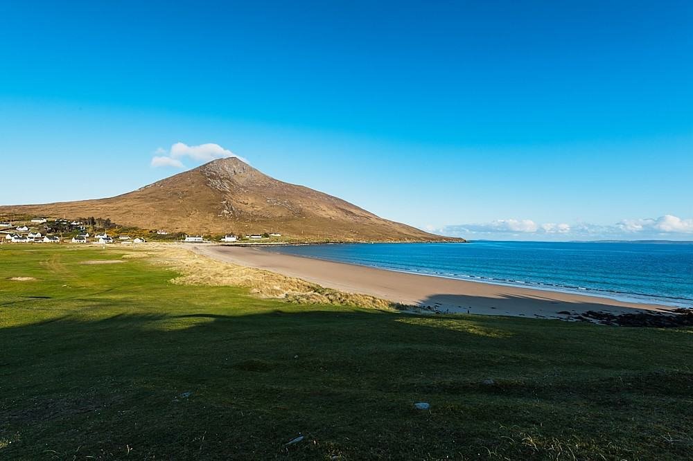 A scenic coastal landscape with a sandy beach, grassy foreground, and a mountain in the background under a clear blue sky.