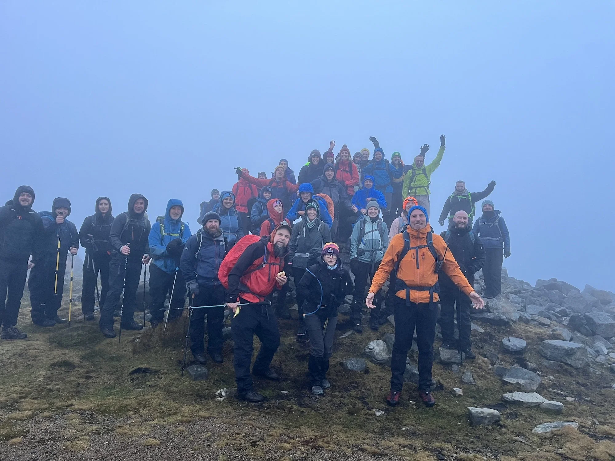 A large group of hikers dressed in outdoor gear and backpacks, standing together on rocky terrain in a foggy mountain setting, some waving and smiling at the camera.