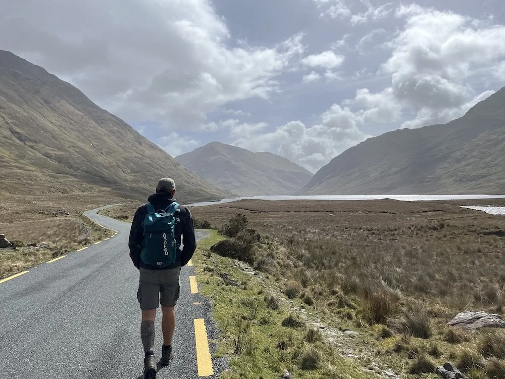A person wearing a backpack walks along a winding mountain road through a scenic valley with mountains on either side and a cloudy sky overhead.