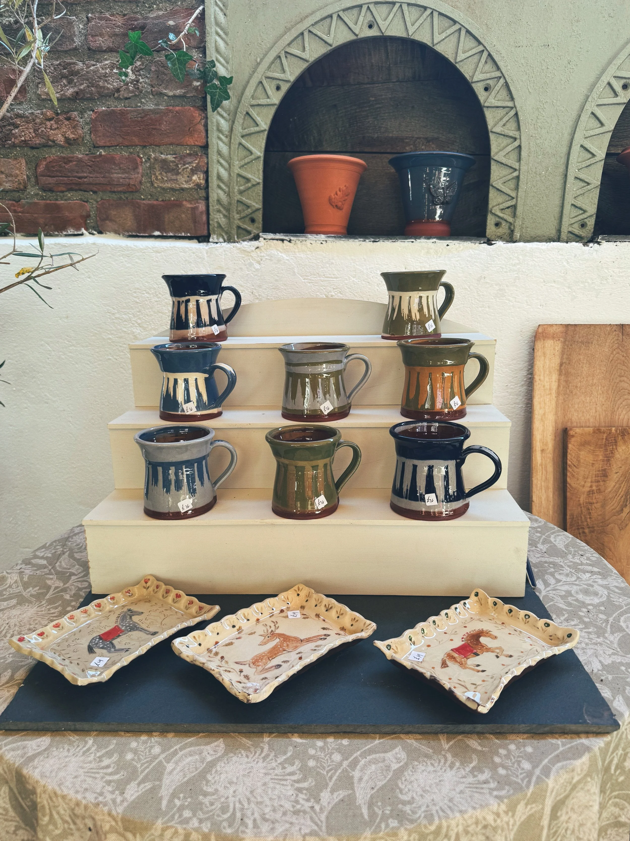 Display of handcrafted pottery including nine glazed ceramic mugs with drip patterns in colors black, green, and blue, arranged on a multi-tiered stand. In front are three decorative ceramic trays with painted animals and festive motifs. In the backg