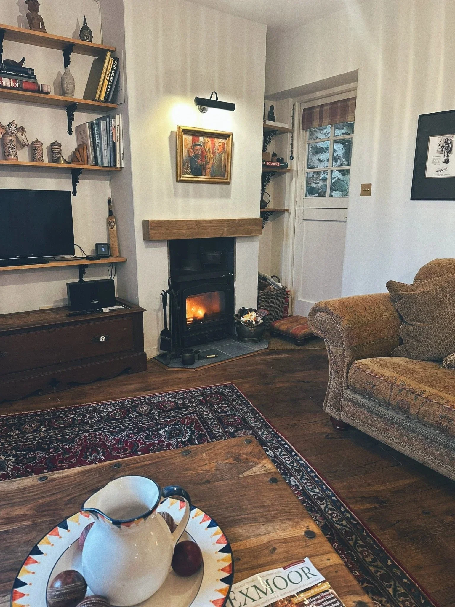 A cozy living room with a wood-burning stove, a patterned rug, a sofa, a wooden coffee table with a decorative pitcher and apples, a bookshelf, a television, and various decorations.