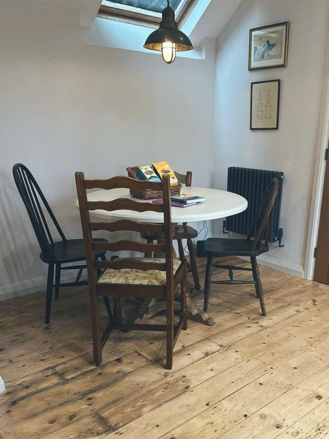 A round white dining table with four chairs, three wooden and one black, in a room with wooden flooring. There is a basket and some items on the table, and a light fixture hanging above. The room has white walls, framed pictures on one wall, and a ra
