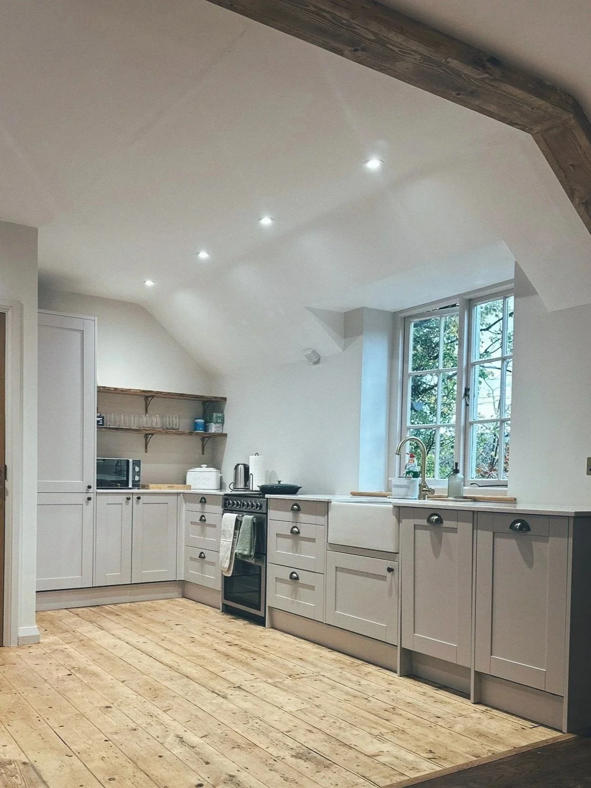 A kitchen with white cabinets, a large window, wooden flooring, and recessed ceiling lights.