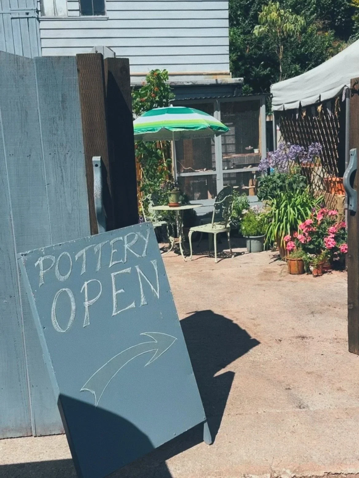 An outdoor pottery shop with a sign that says 'Pottery Open' and an arrow pointing to the right. There are outdoor tables, chairs, a green umbrella, and potted plants and flowers.