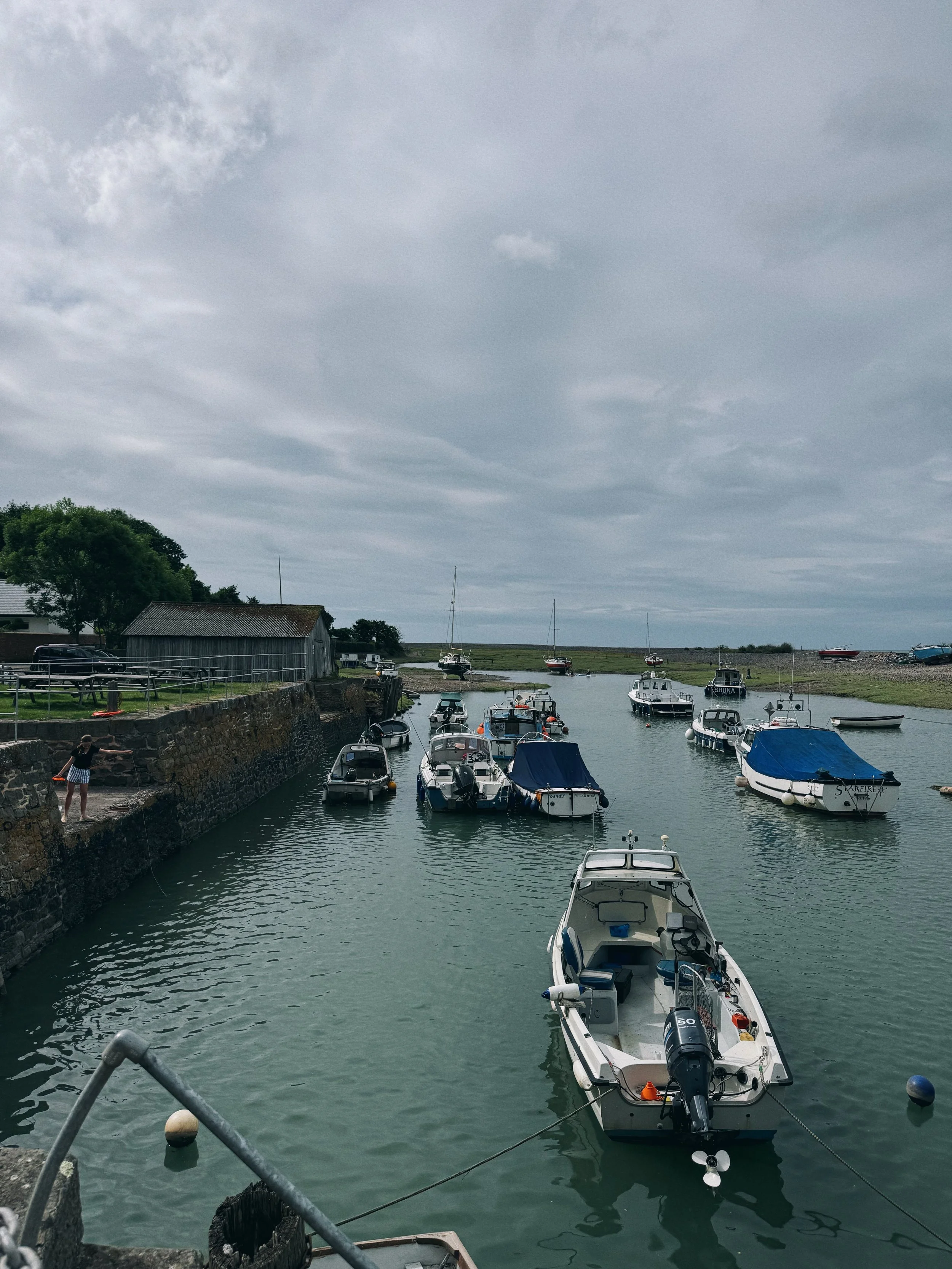 Boats moored in a small marina with overcast sky and a dock with a person fishing.