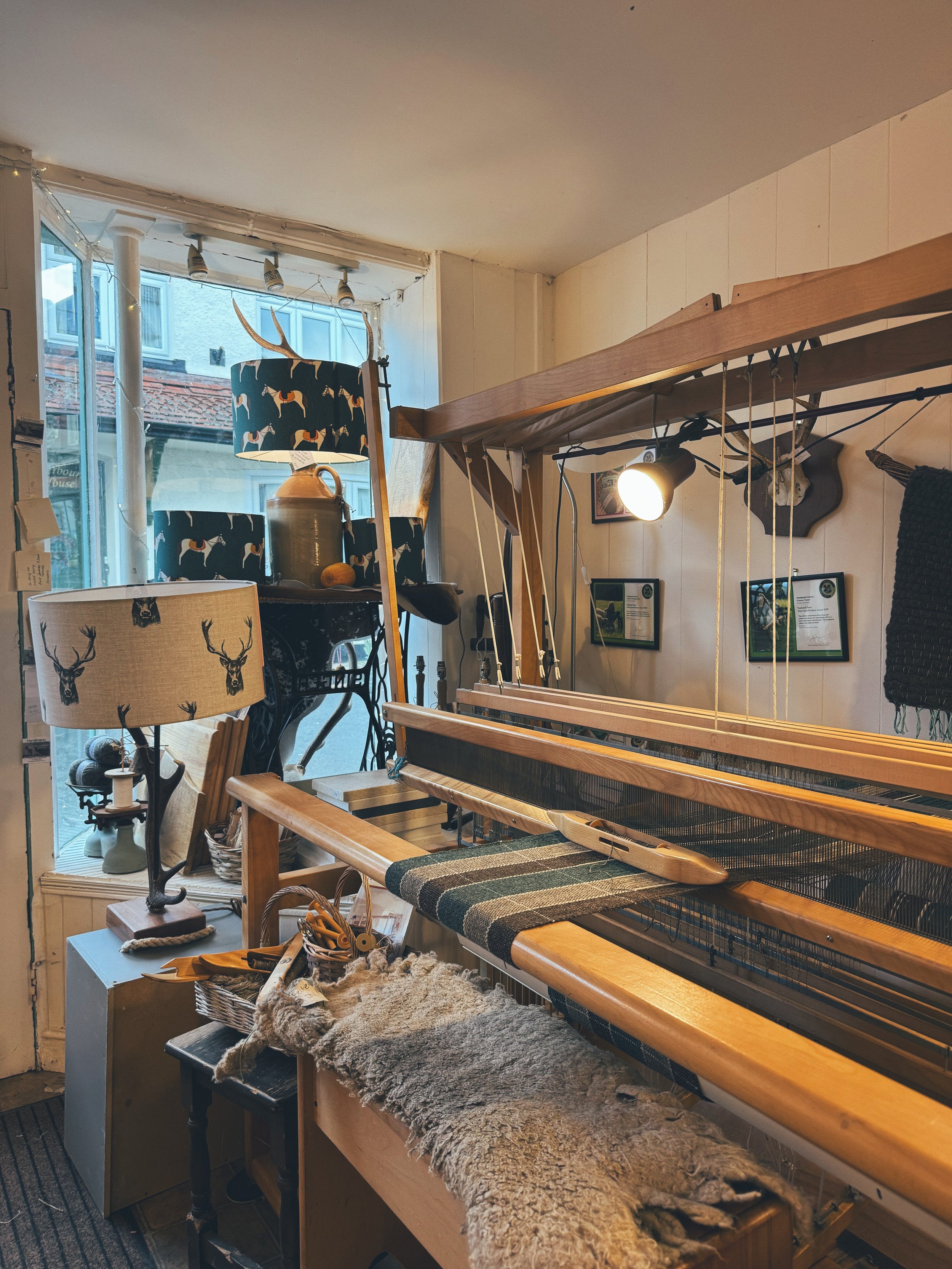 Interior of a craft or weaving shop with a loom, decorative lampshades, and various woven and crafting items, illuminated by natural light through a window.