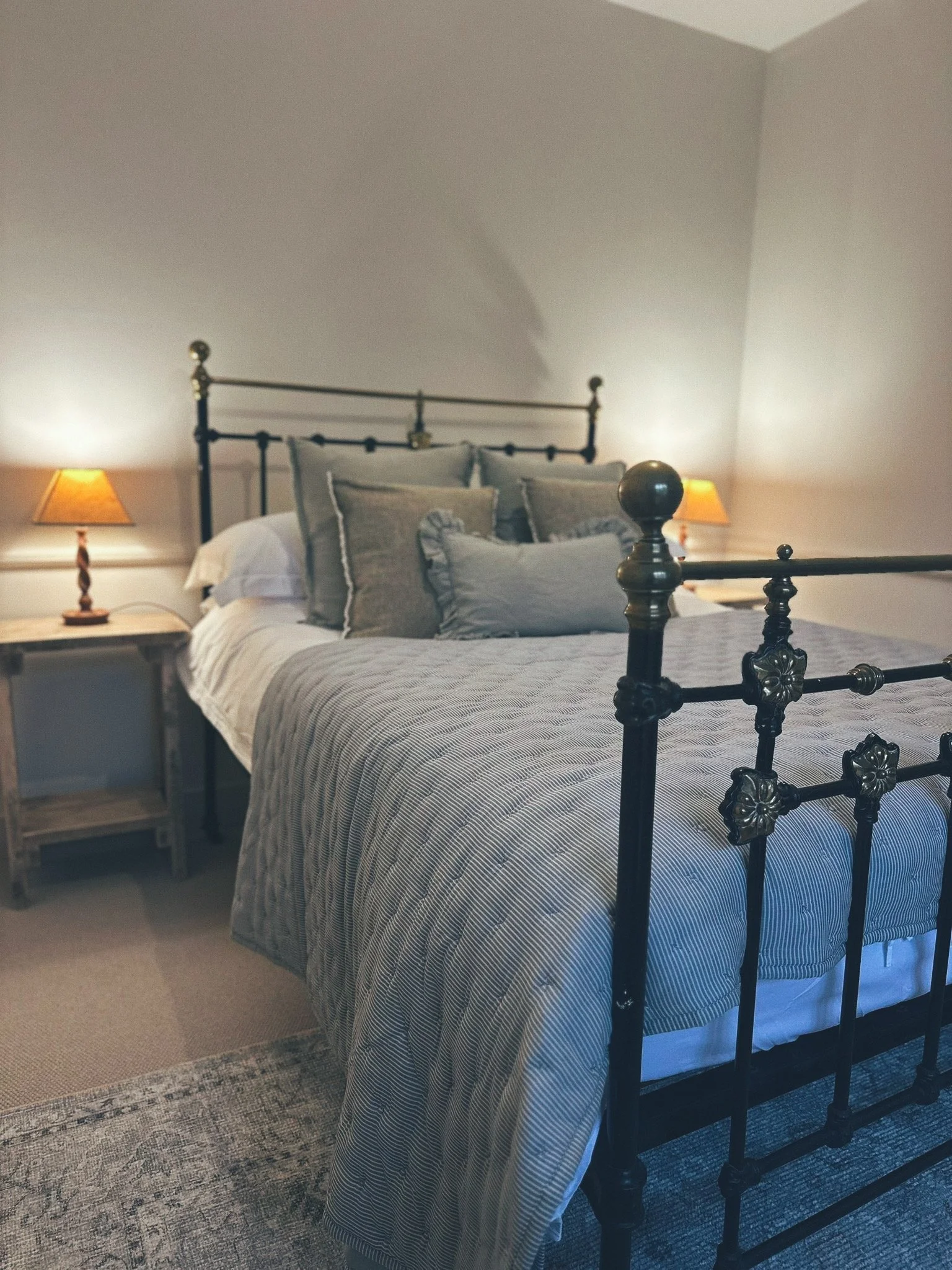 A bedroom with a black metal bed frame, light-colored bedding, several pillows, and two bedside lamps with orange shades in a softly lit room.