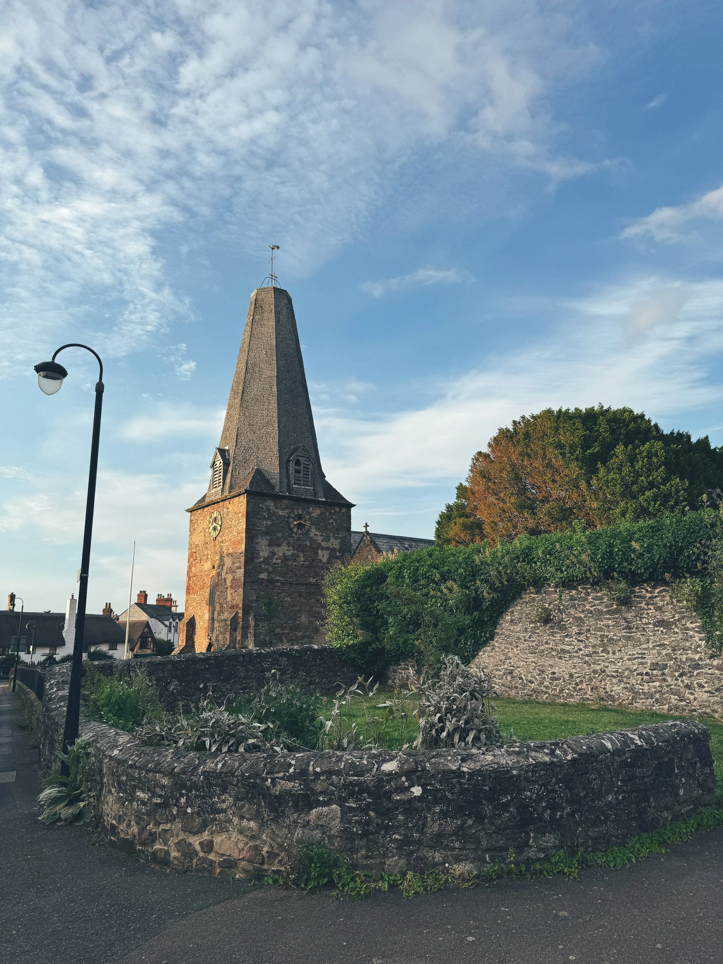 A historic stone church with a tall, pointed steeple and a weather vane on top, adjacent to a stone wall and lush greenery under a blue sky with scattered clouds.