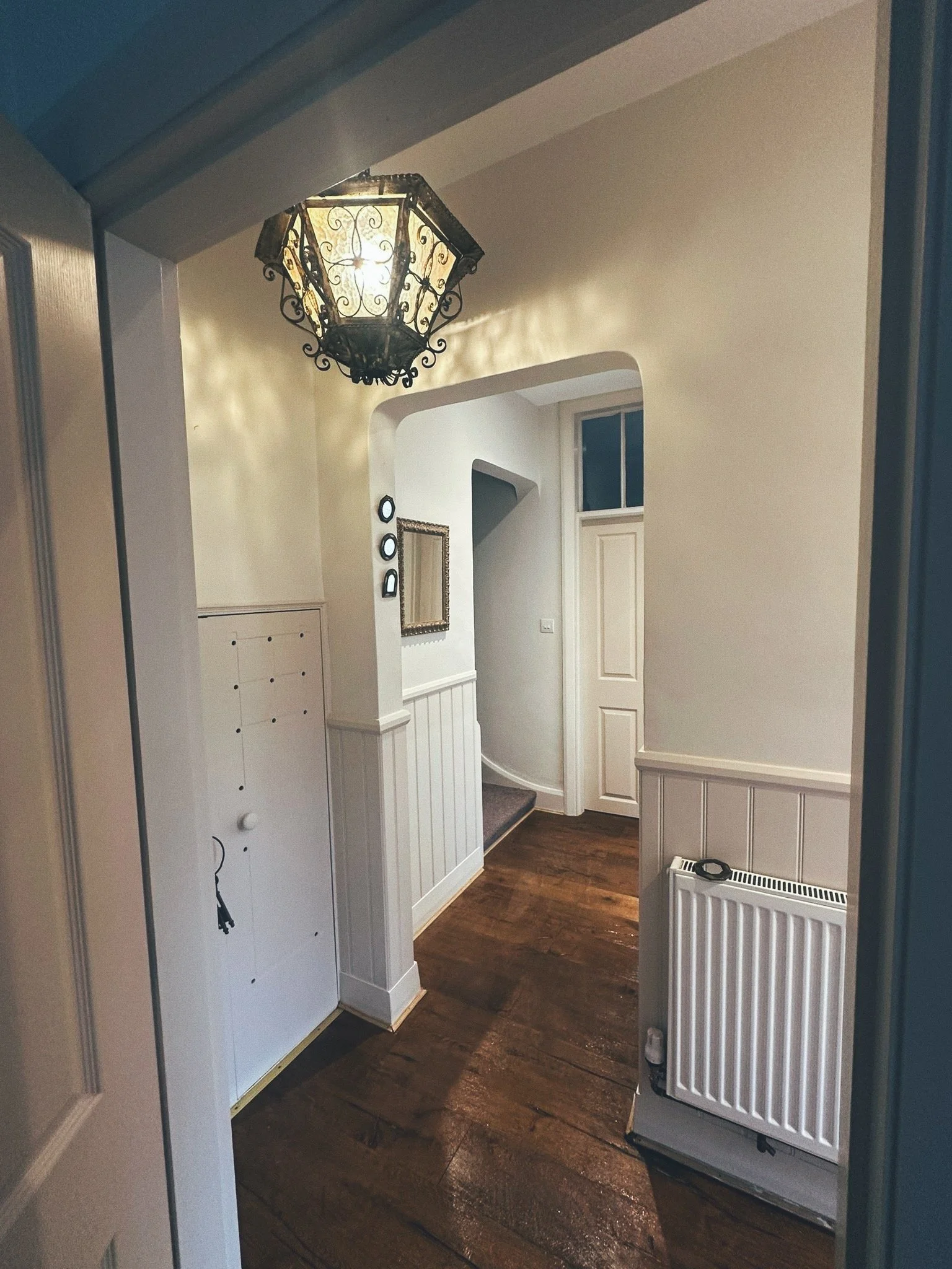 Interior view of a house entryway with hardwood floors, white walls with paneling, a decorative black and white lantern ceiling light, a small mirror, a radiator, and stairs leading upward.