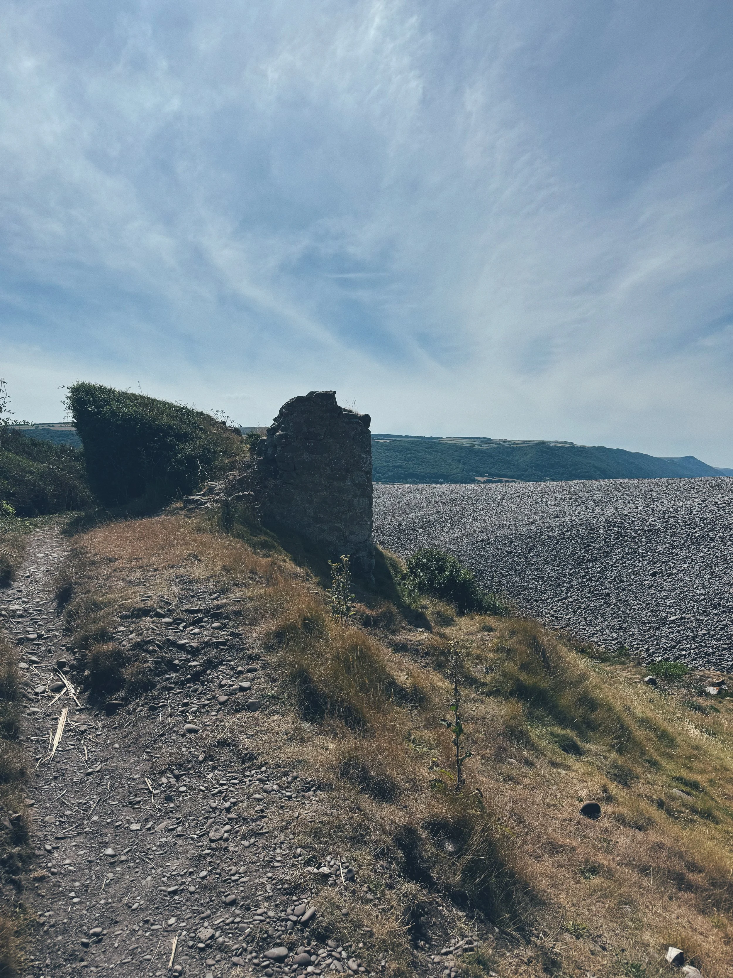 A rugged coastal landscape with a dirt path, a stone structure, grassy terrain, and distant hills under a partly cloudy sky.