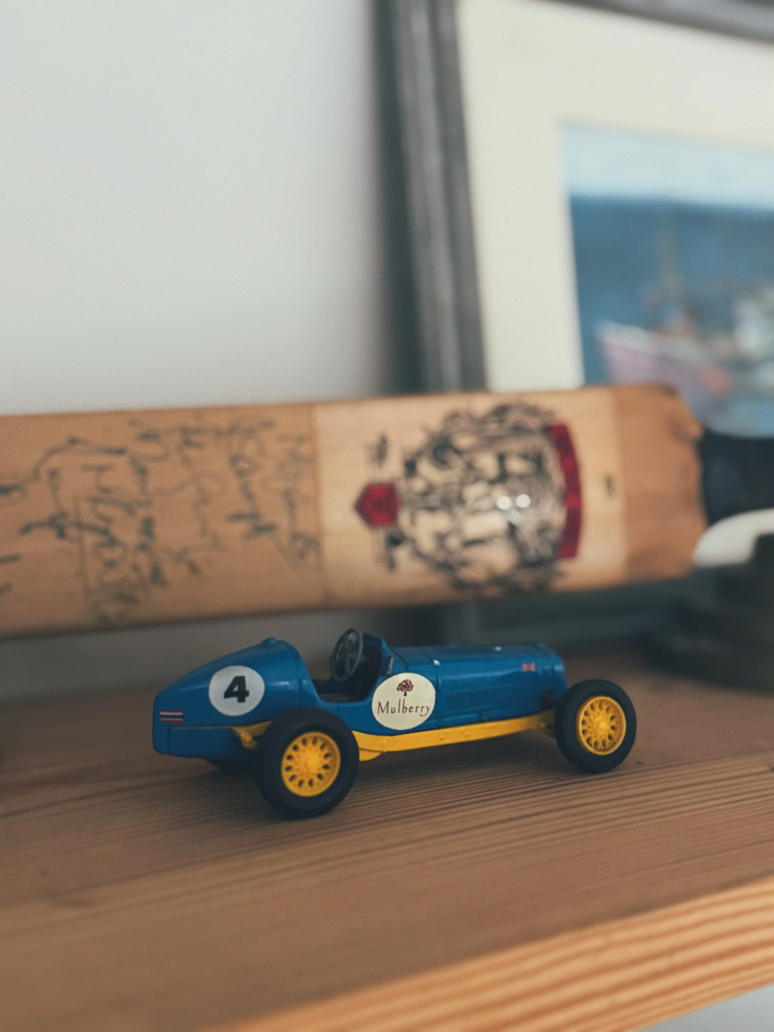Small blue toy race car with yellow wheels and a white circle with the number 4, placed on a wooden surface with a blurred framed picture and a wooden object in the background.