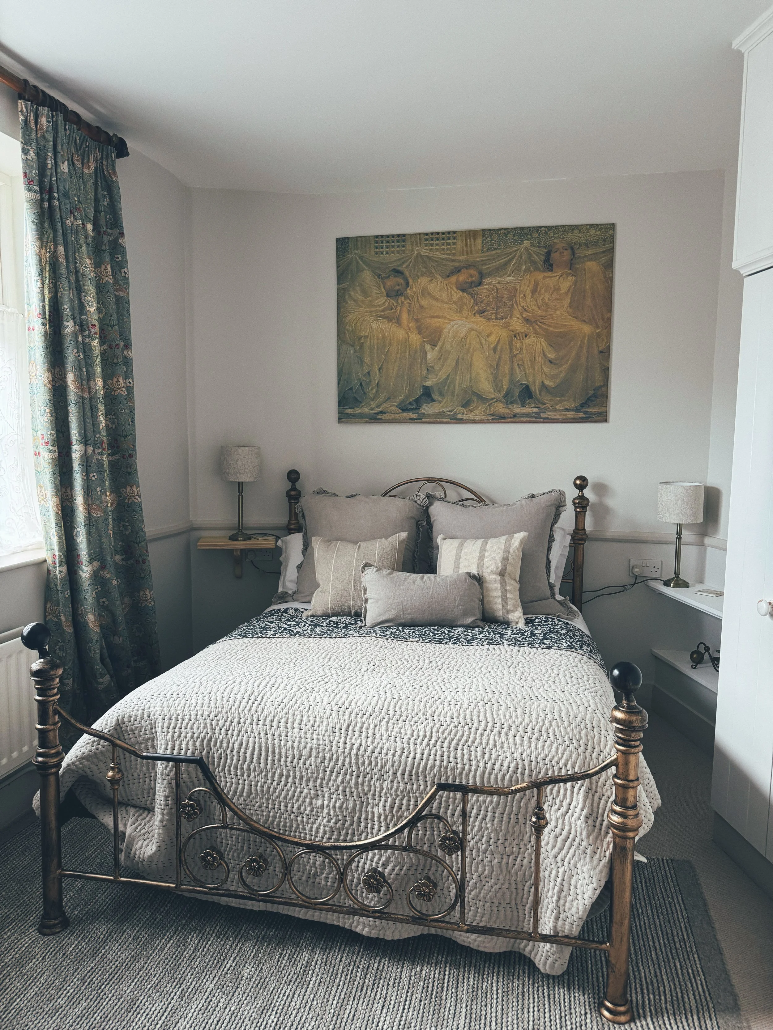 A bedroom with a vintage metal bed, beige and gray bedding, four pillows, a wall-mounted bedside table with a lamp on each side, a framed painting above the bed, a window with floral curtains, and a gray carpet.