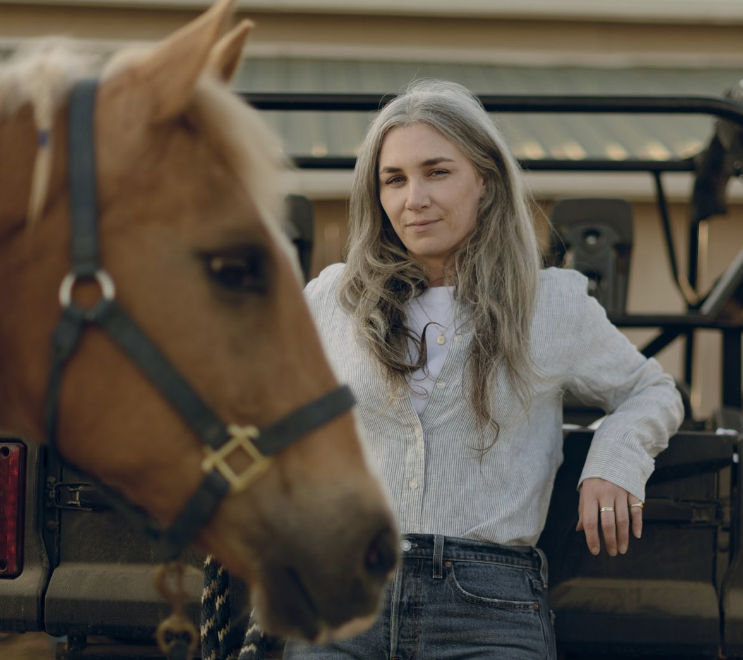 Young woman with long gray hair leaning against a vehicle, gazing at a palomino horse in front of her.