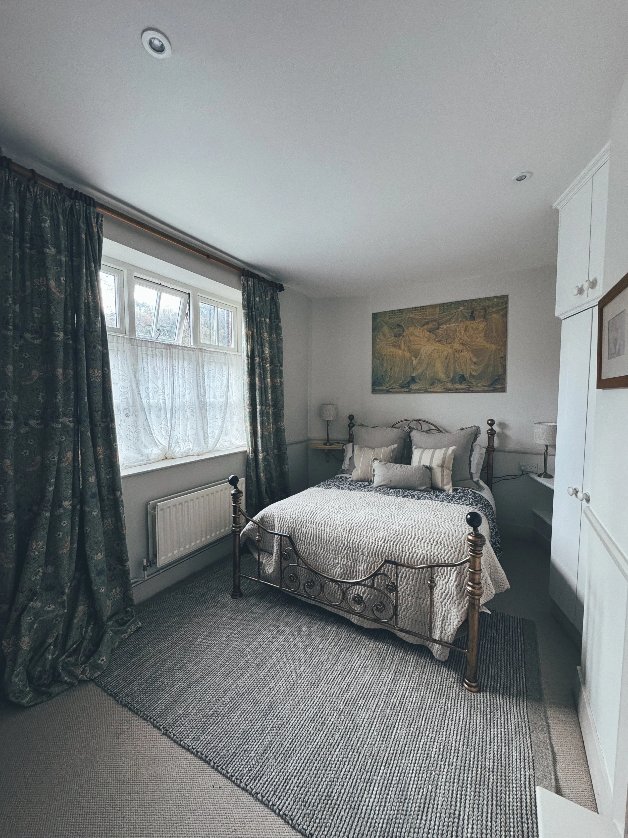 A bedroom with a bed, pillows, and a quilt, a window with curtains, a radiator, a painting above the bed, and a wardrobe.
