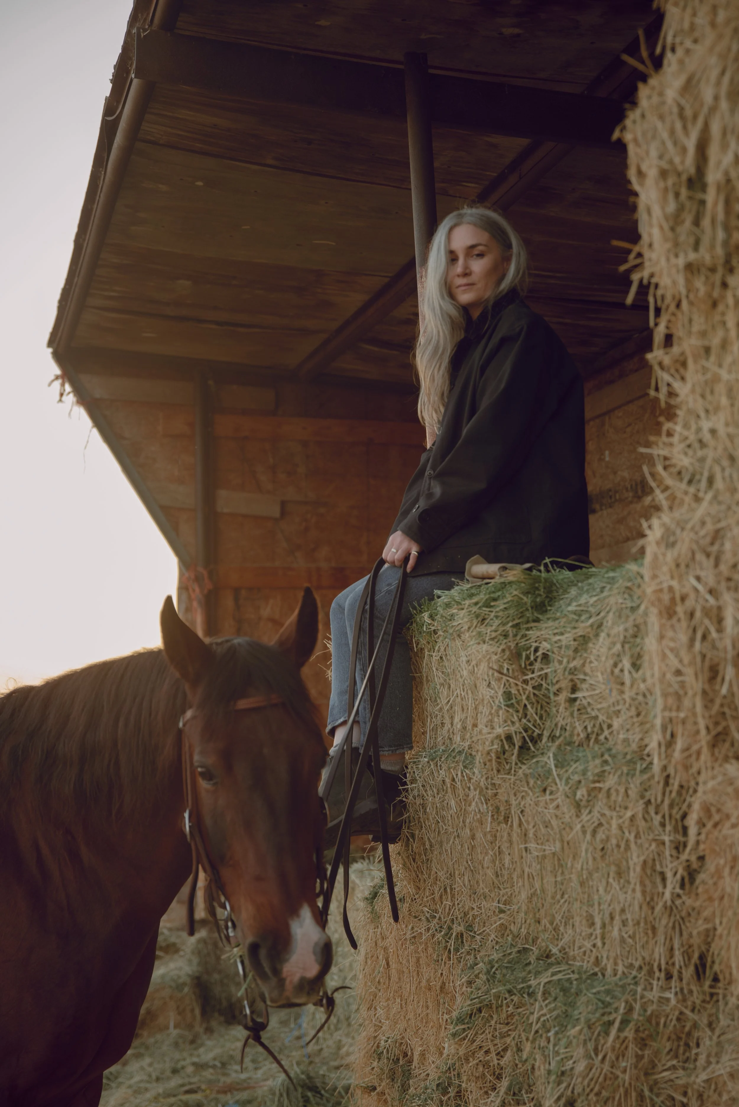 Storme Whitby-Grubb wearing Filson overshirt sitting on hay bails with her sorrel horse, Sprocket