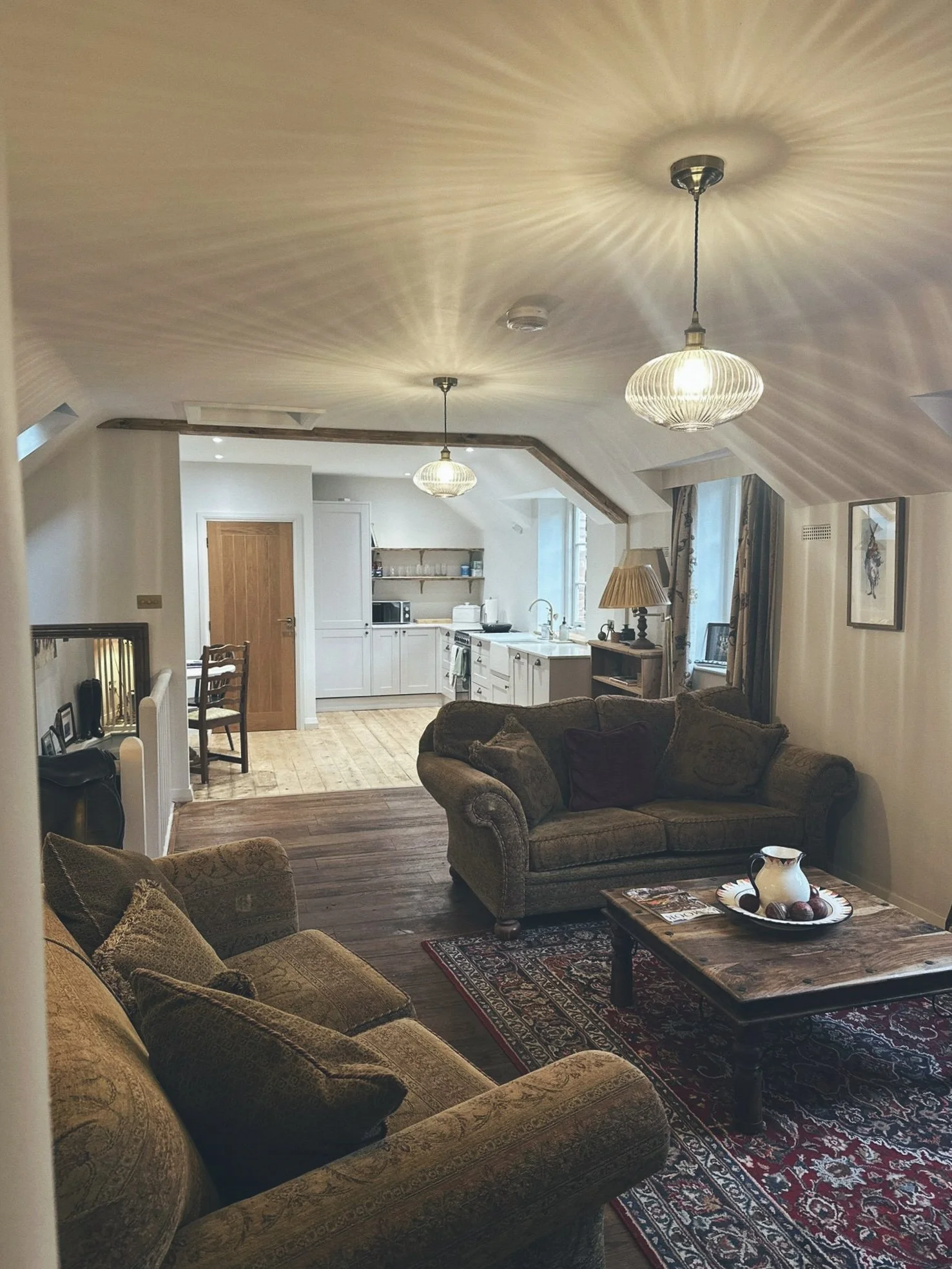 Living room with sofas, a wooden coffee table, and a rug, leading to an open kitchen with white cabinets and a window.