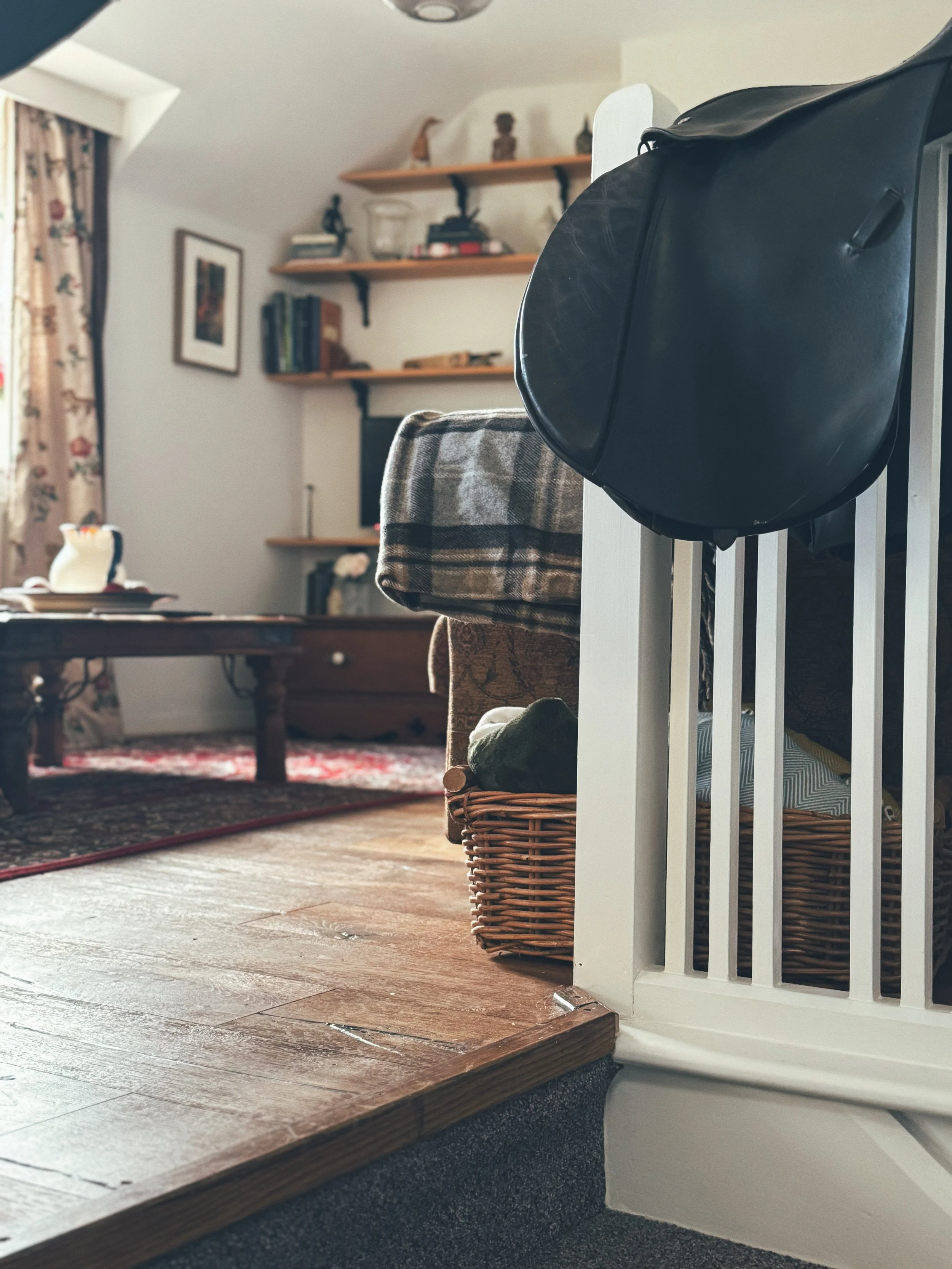 Interior view of a living room showing a plaid blanket draped over a piece of furniture, a wicker basket with some items inside, a black bag hanging on a white railing, and a wooden dining table with books and a teapot in the background. The room has