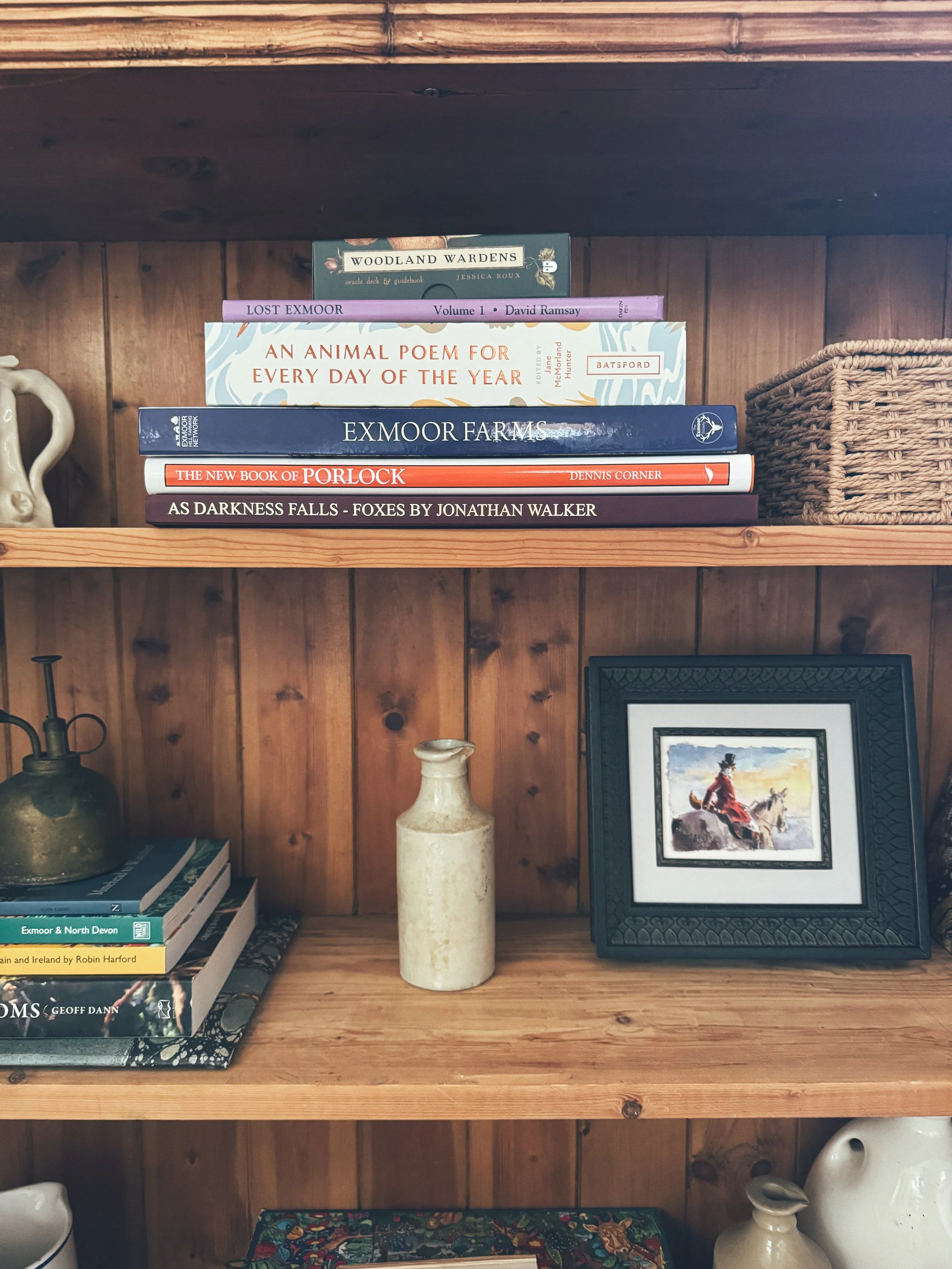 Wooden bookshelf with books, framed watercolor painting of a rider on a horse, ceramic vases, and a woven basket.