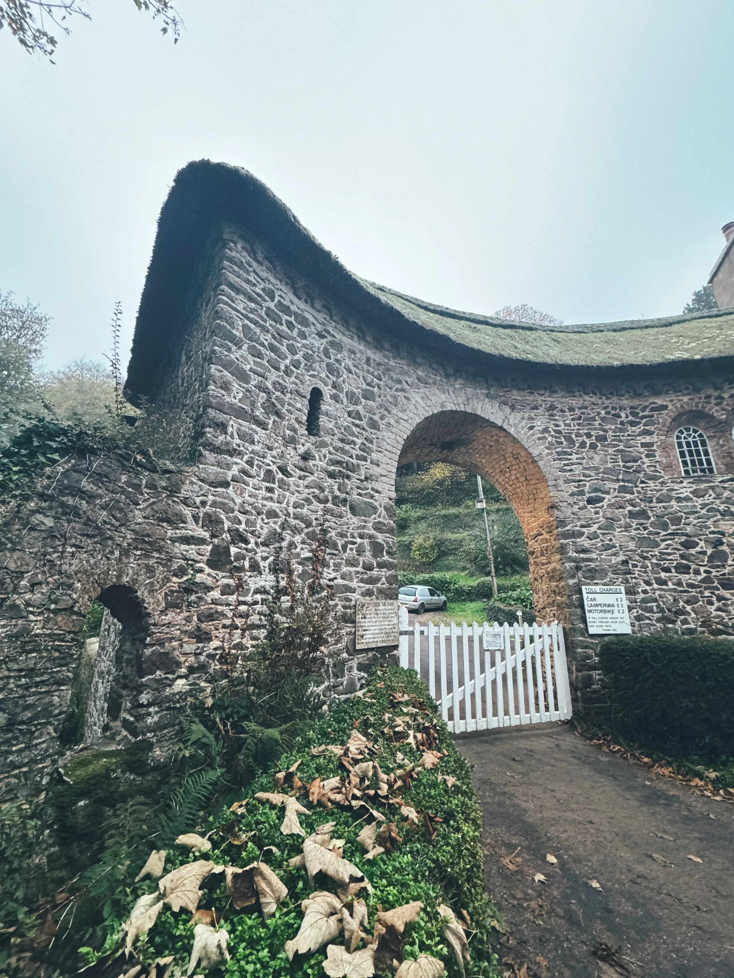 Stone archway entrance to a historic building with a thatched roof, a white gate, and signs with toll charges, surrounded by greenery and fallen leaves.