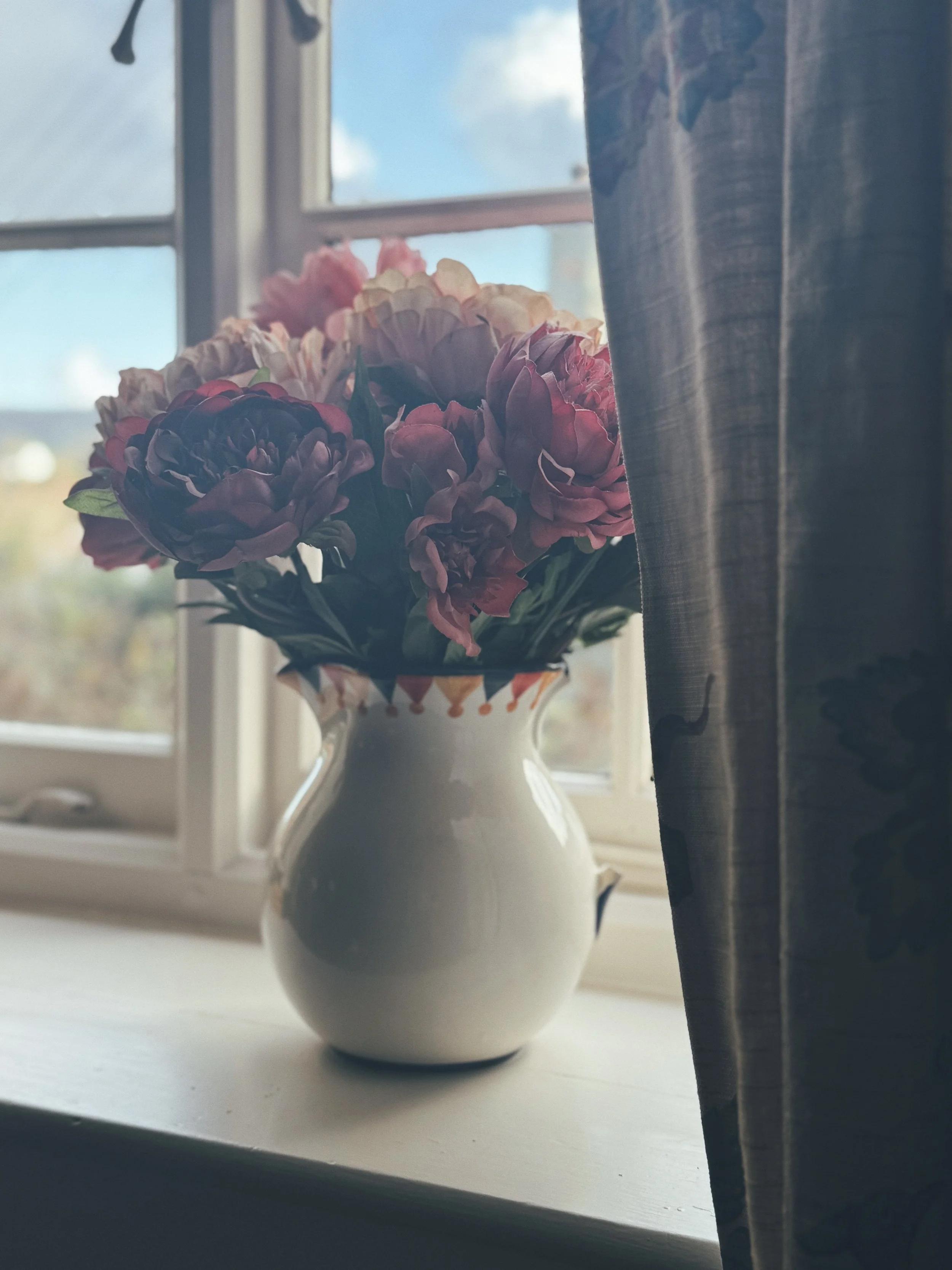 A vase with pink and purple flowers sitting on a windowsill beside a curtain, with a partly cloudy sky visible through the window.