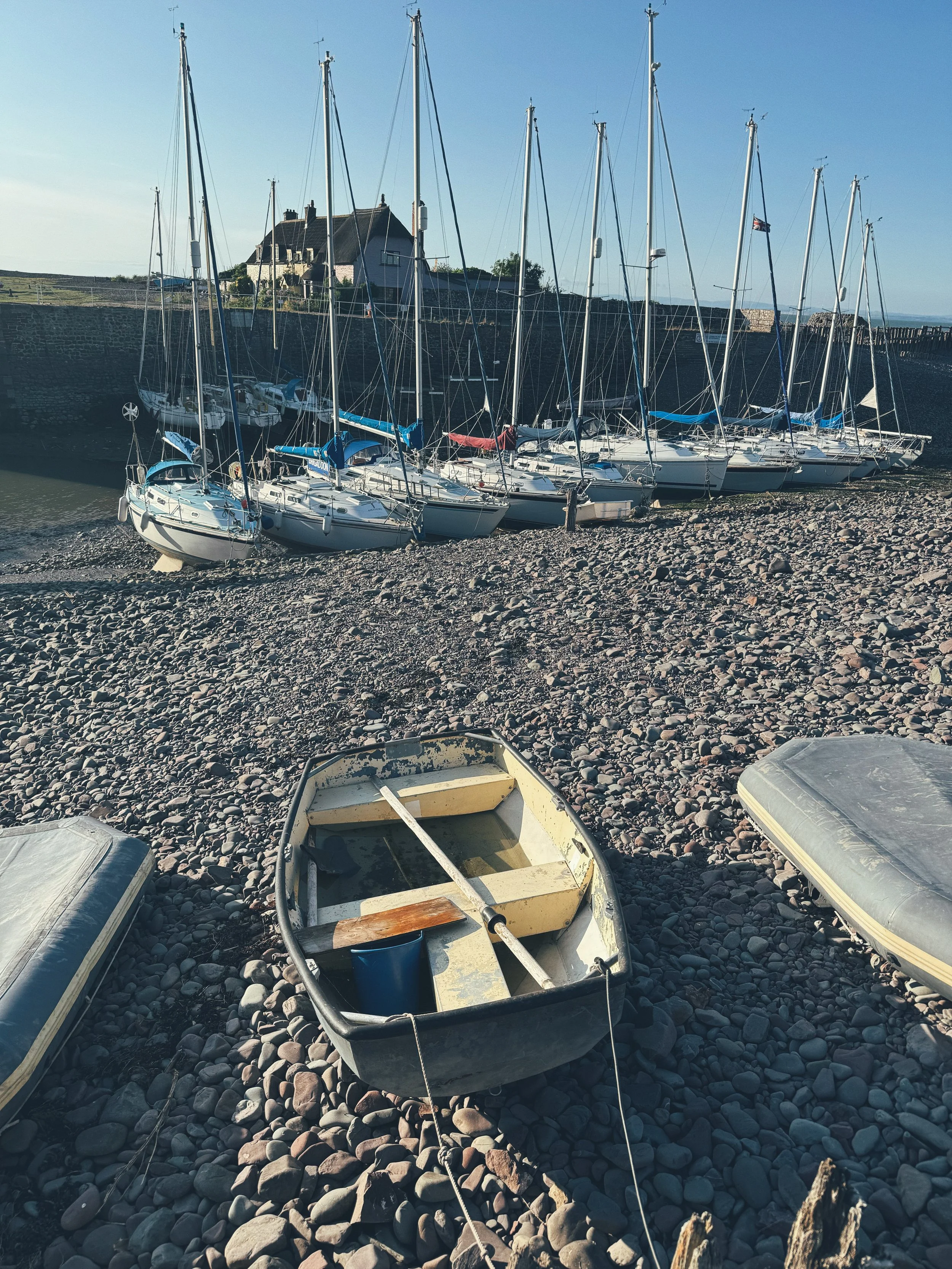 A small rowboat resting on a rocky shore in front of a marina with several sailboats docked.