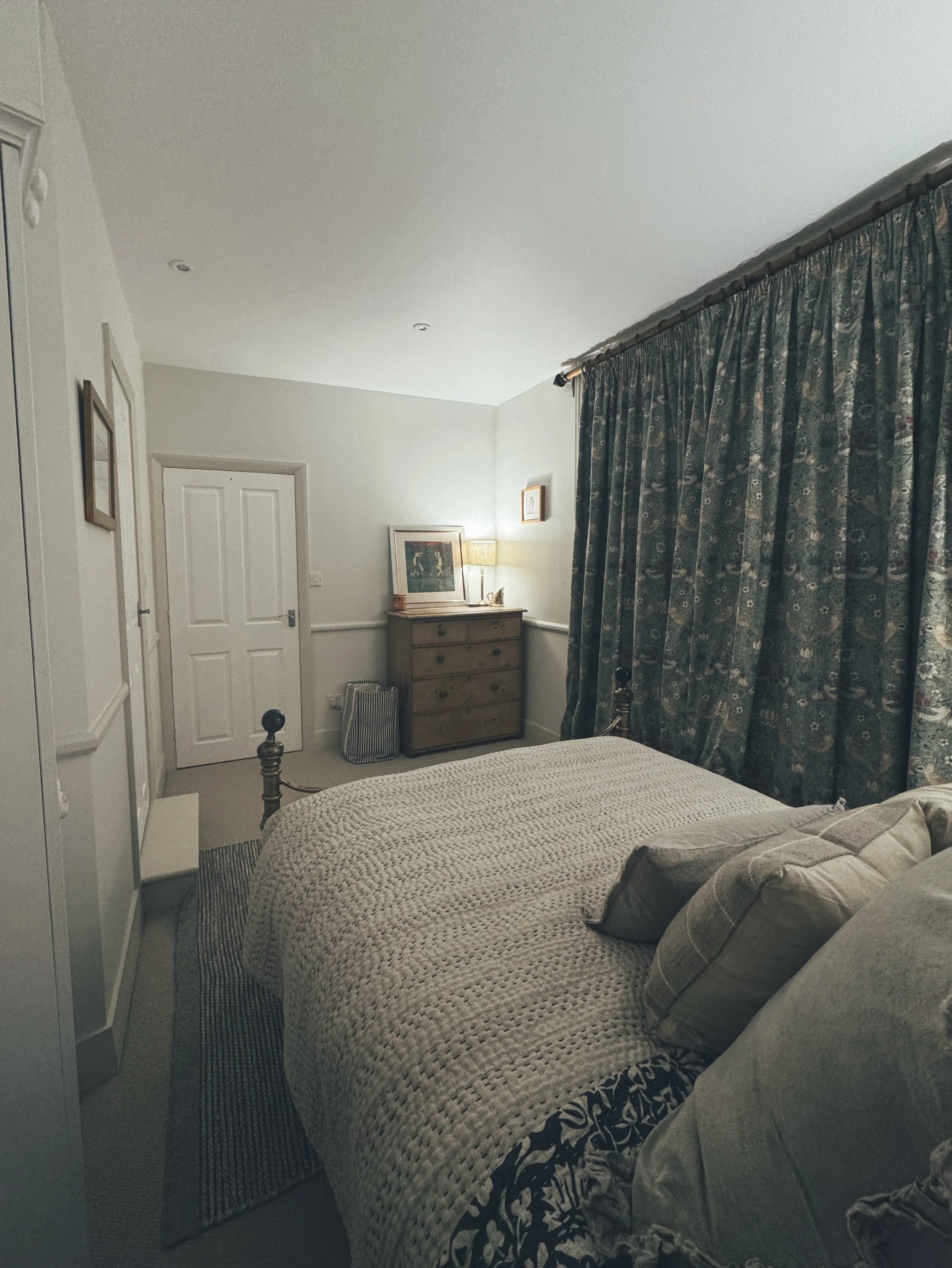 A cozy bedroom with a bed covered in a beige knitted blanket and pillows, a wooden dresser with a lamp and framed artwork, and dark patterned curtains over the window.