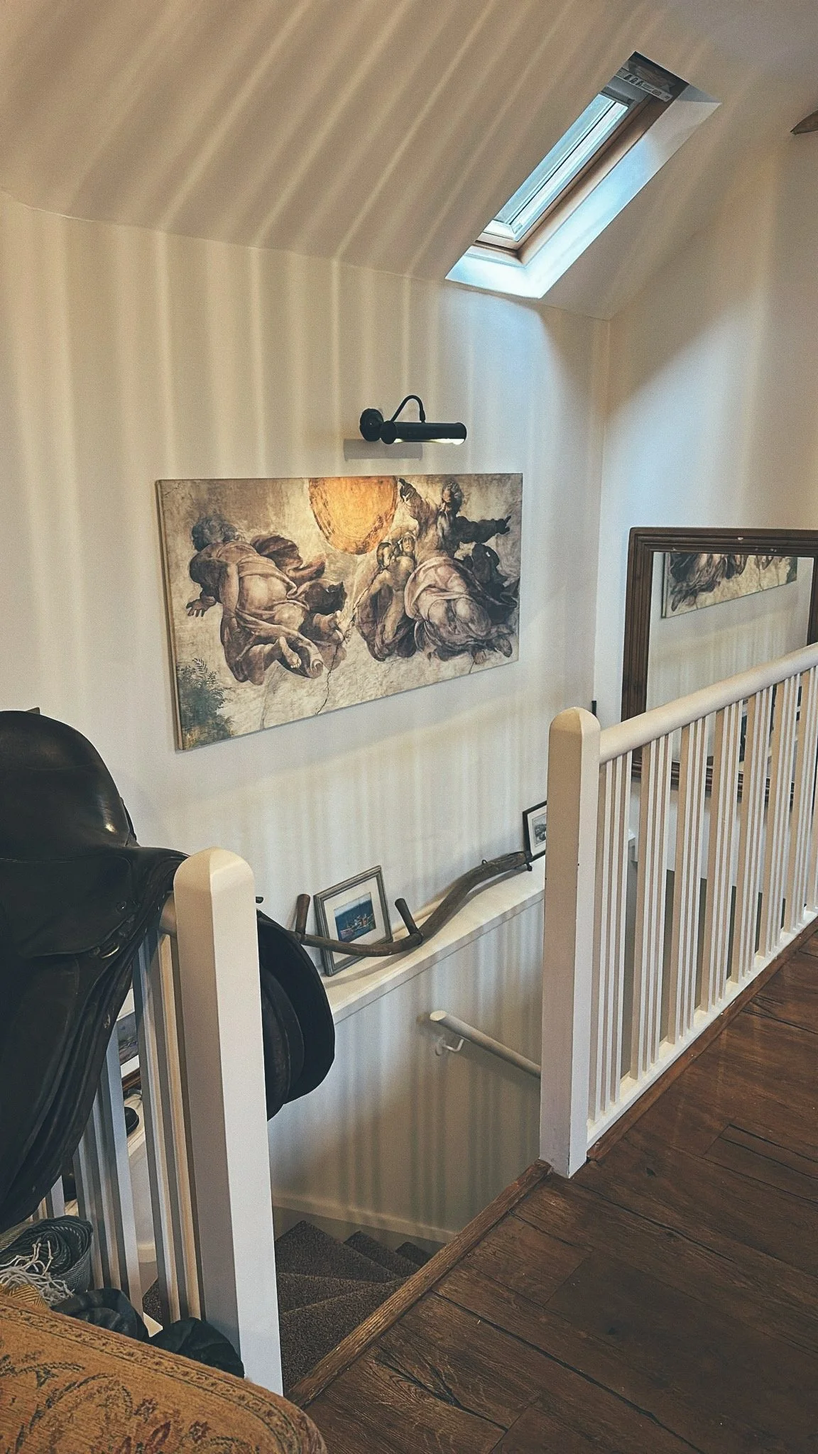 An indoor staircase with a wooden handrail and carpeted steps, leading down to a lower level. The wall on the upper level features artwork, framed photos, and a mounted camera or light fixture. There is a skylight window on the sloped ceiling bringin