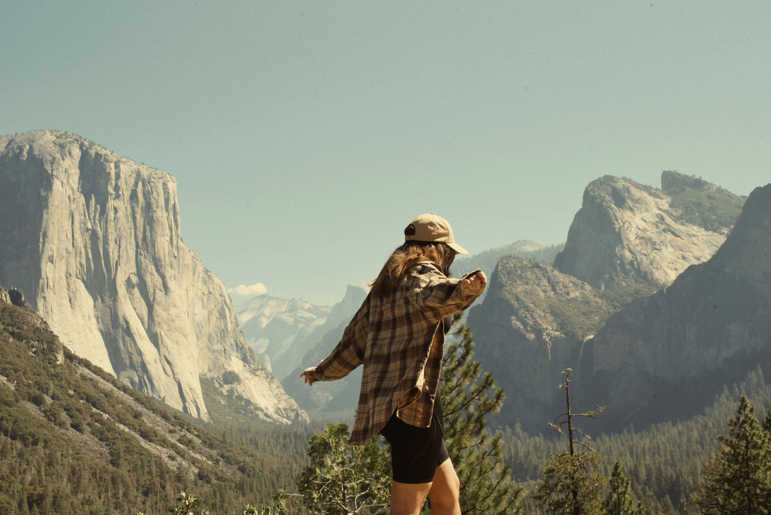 A woman in hiking attire, including a cap and a plaid shirt, standing in front of a mountainous landscape with large rock formations and a clear sky.