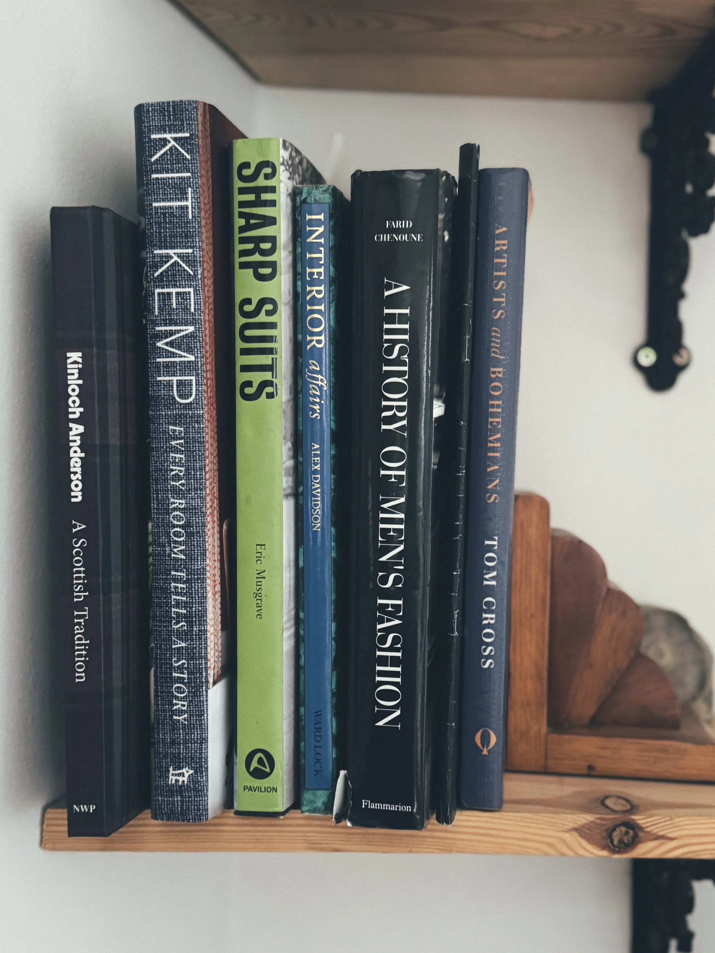 A wooden bookshelf holding seven books about history and storytelling, with a wooden wall and decorative black metal element above.