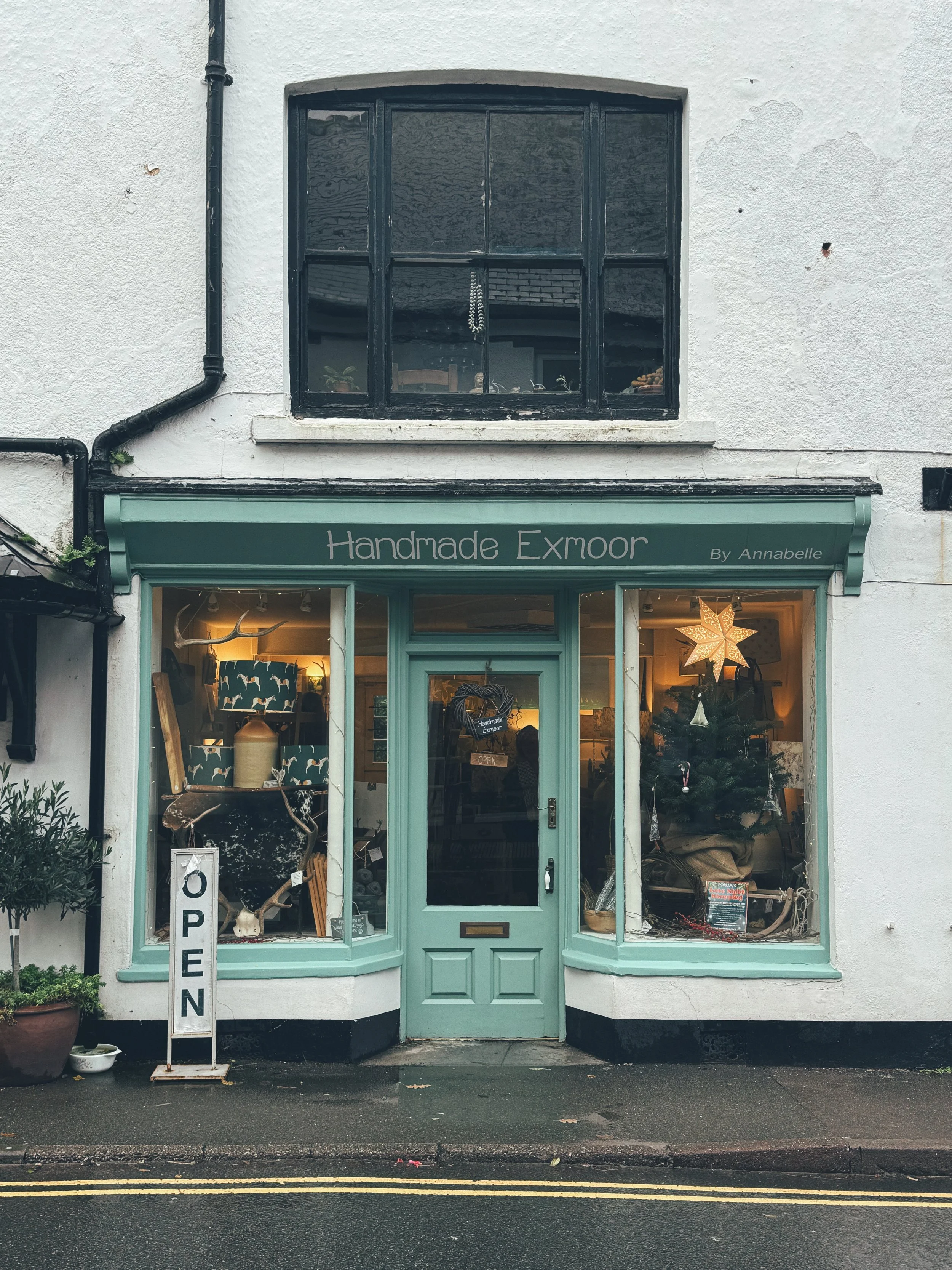 Front view of a small shop named Handmade Exmoor by Annabelle with a light green door and window frames, decorated for Christmas with a star ornament and a small Christmas tree inside. An 'OPEN' sign is displayed outside.