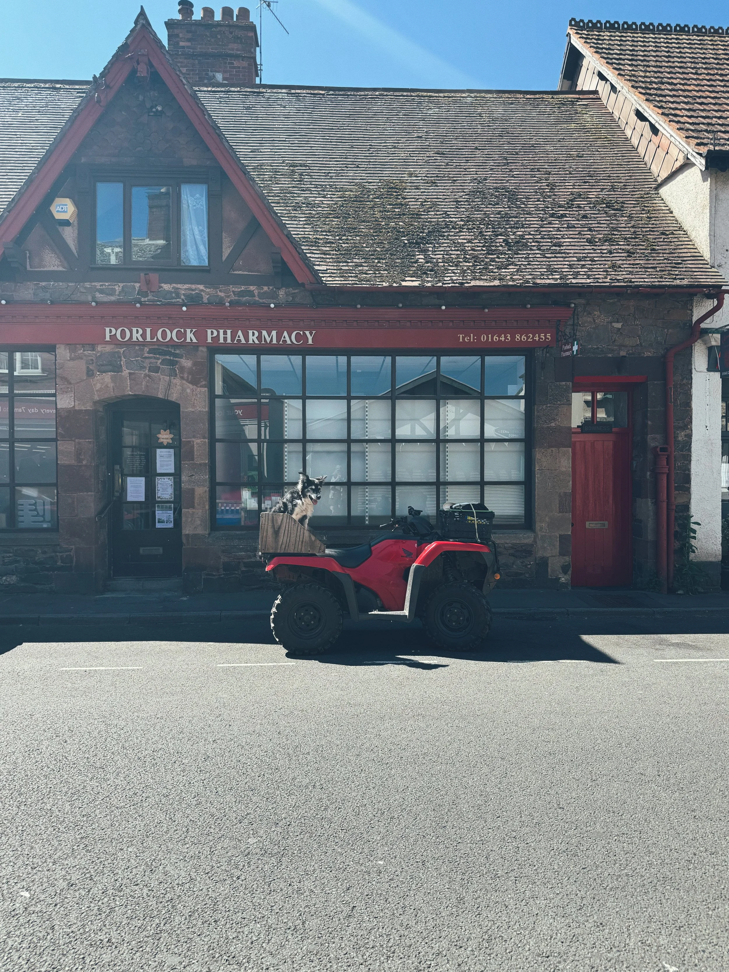 A small brick building with a sign reading "Porlock Pharmacy" on the front, situated on a street. A red ATV is parked in front of the building, with a dog sitting on it.