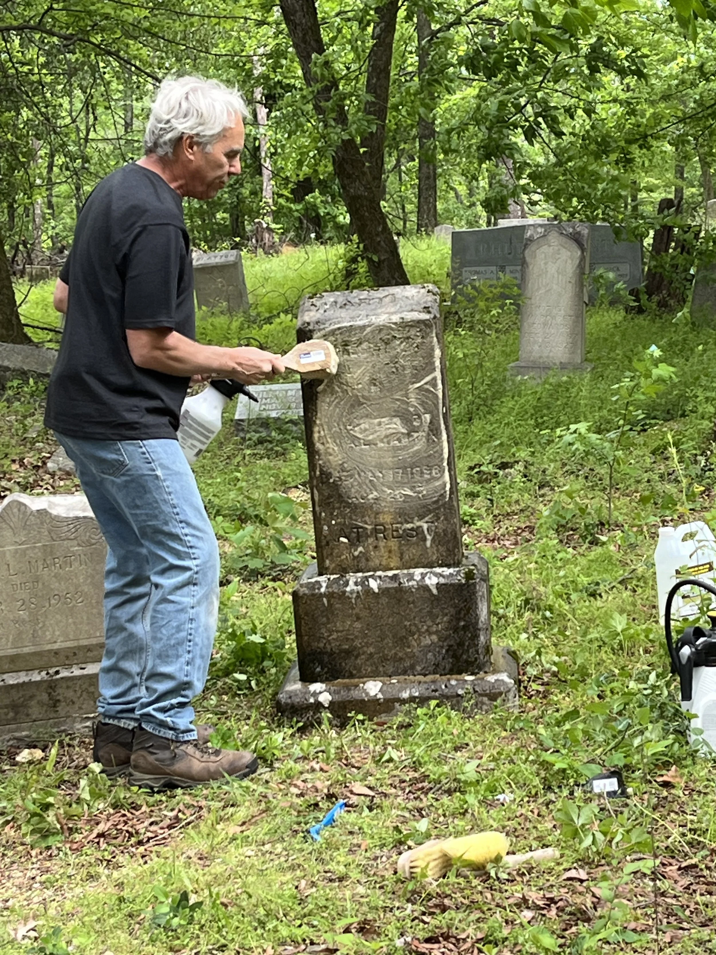 A man cleaning a weathered tombstone in a green, wooded cemetery.