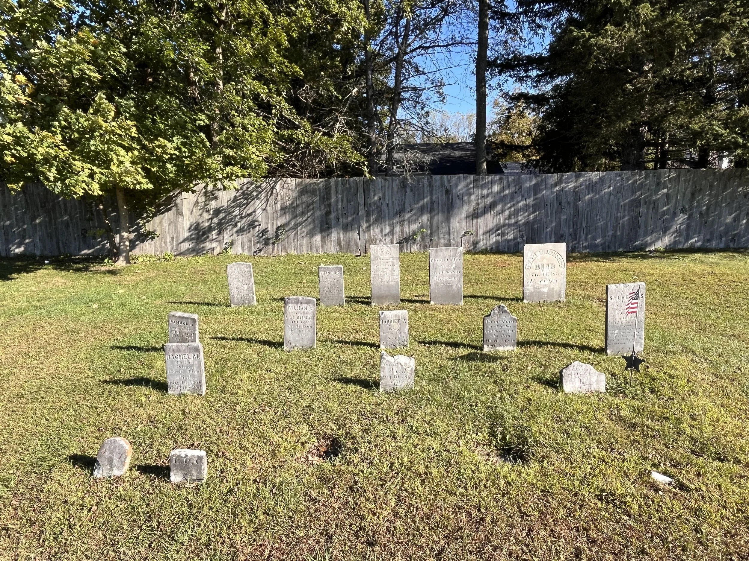 Cemetery with small worn tombstones arranged in rows on grass, surrounded by trees and a wooden fence.