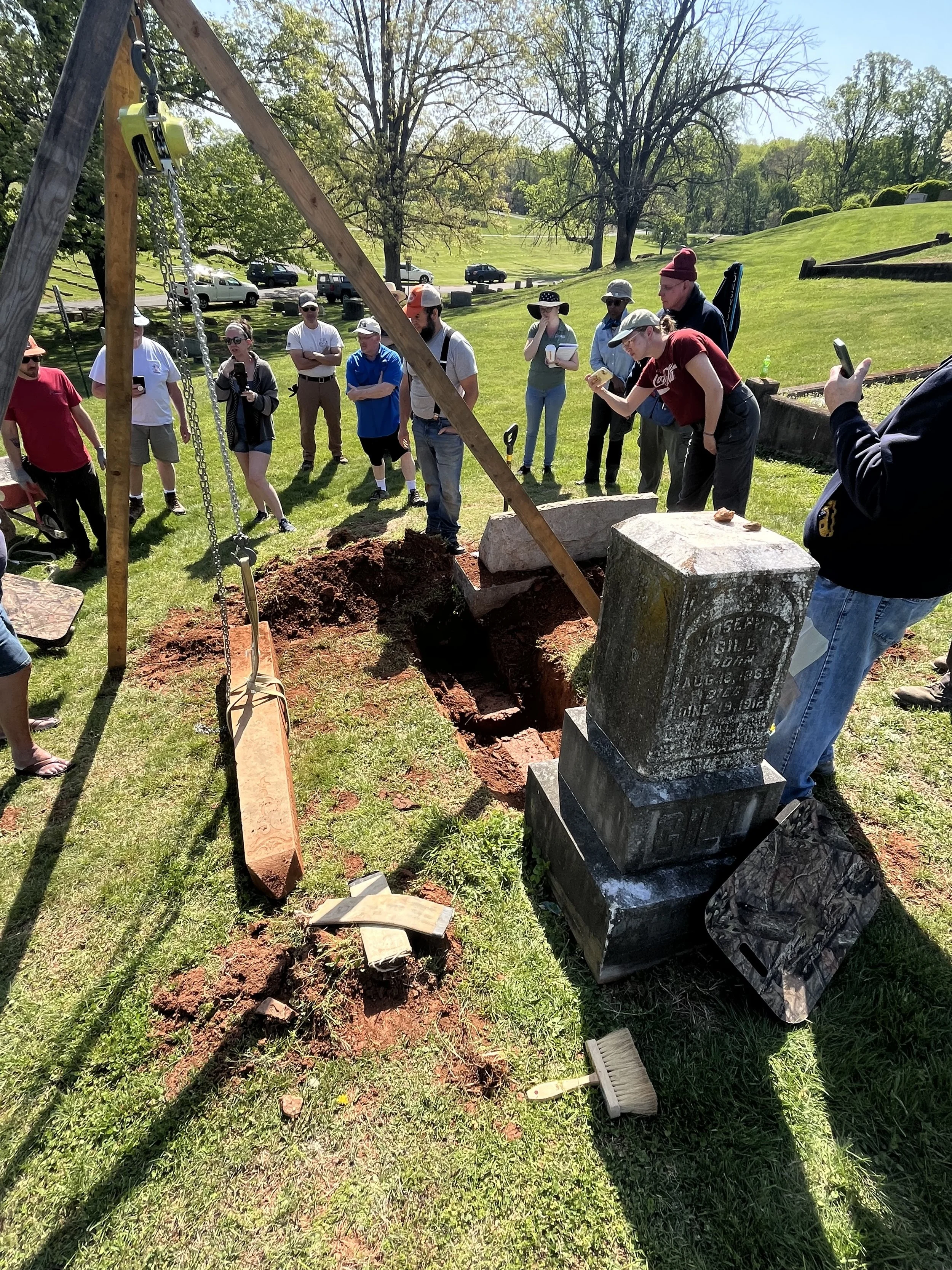 A group of people gathered around a grave site with a fresh excavation in a cemetery, with trees and a grassy landscape in the background.