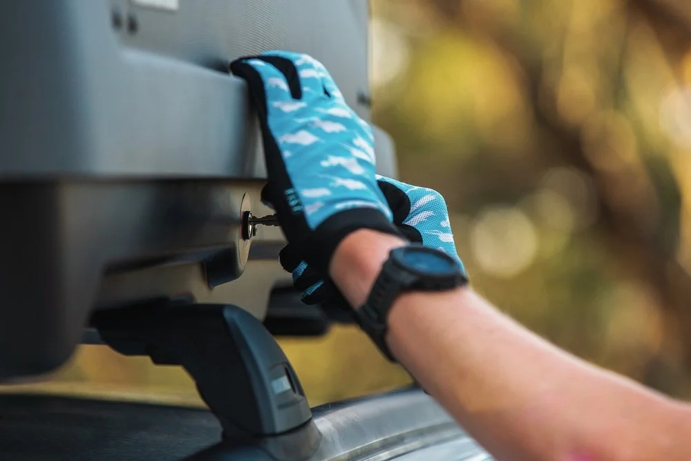 A person wearing a black watch and blue gloves is inserting a key into a lock on a black metal trunk or toolbox outdoors with blurred trees in the background.
