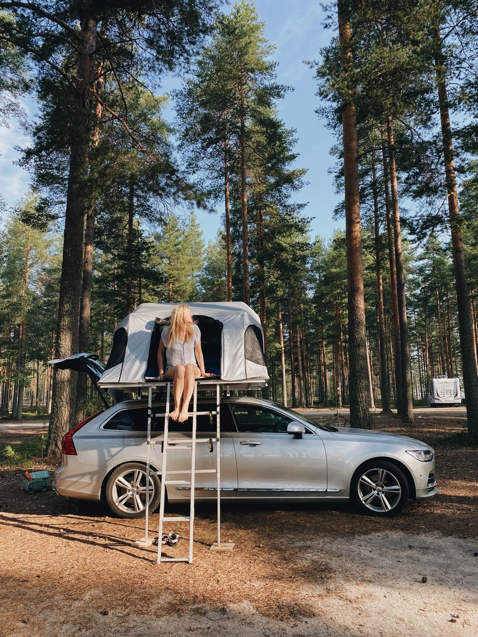 A woman sitting in a rooftop tent on top of a silver car in a forest with tall pine trees and a clear blue sky.