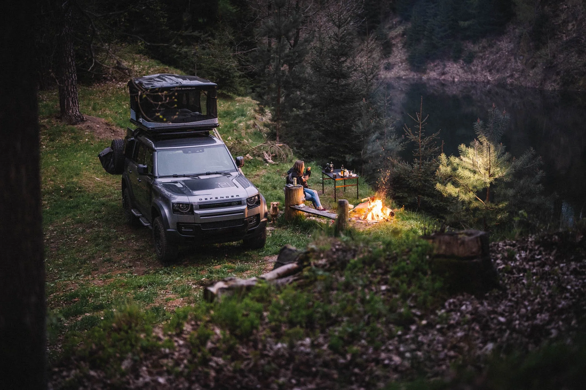 A woman sitting by a campfire near a lake, with a black SUV parked nearby and a table with drinks and supplies, surrounded by trees at dusk.