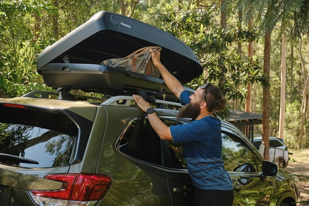 A man loading a suitcase into a roof cargo box on a dark green SUV in a wooded outdoor area.