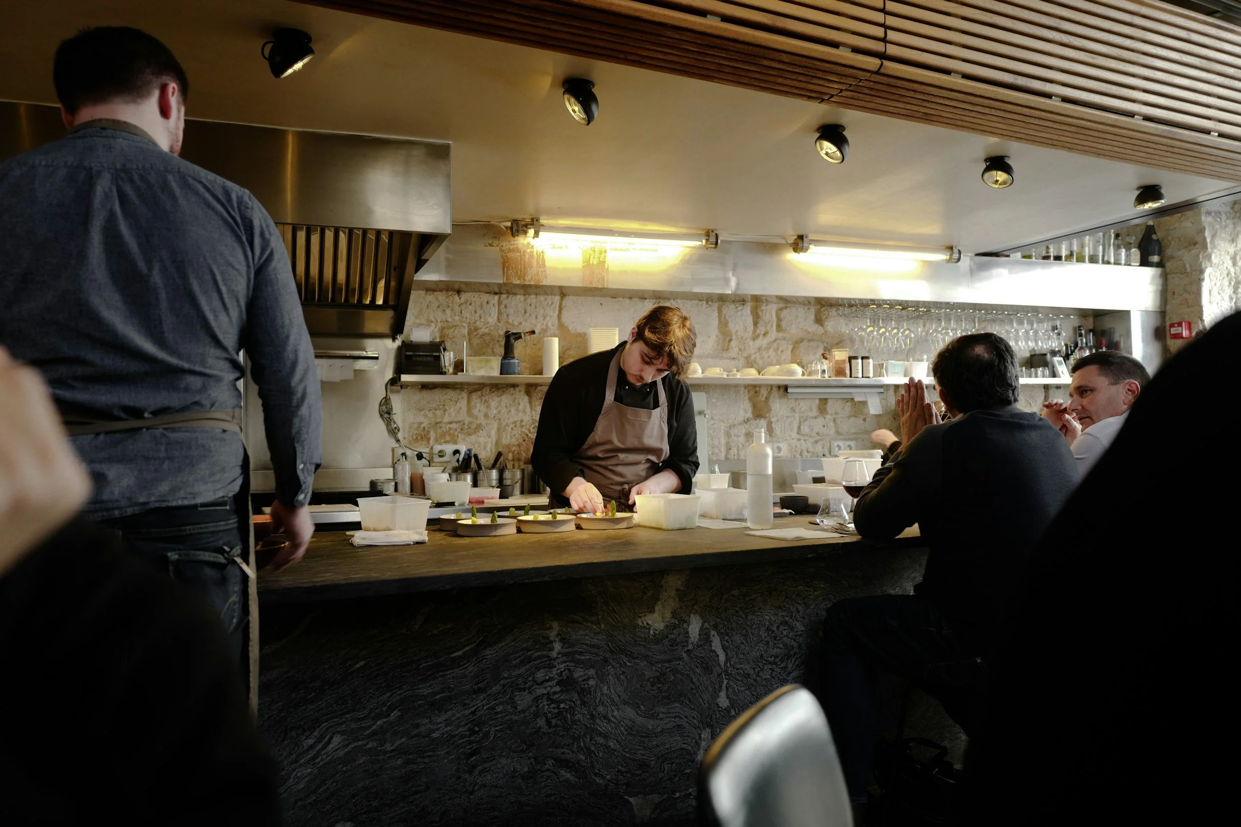 A chef preparing food at a restaurant kitchen while diners are seated at a counter watching. The kitchen has a rustic stone wall, shelves with glasses, and modern lighting.