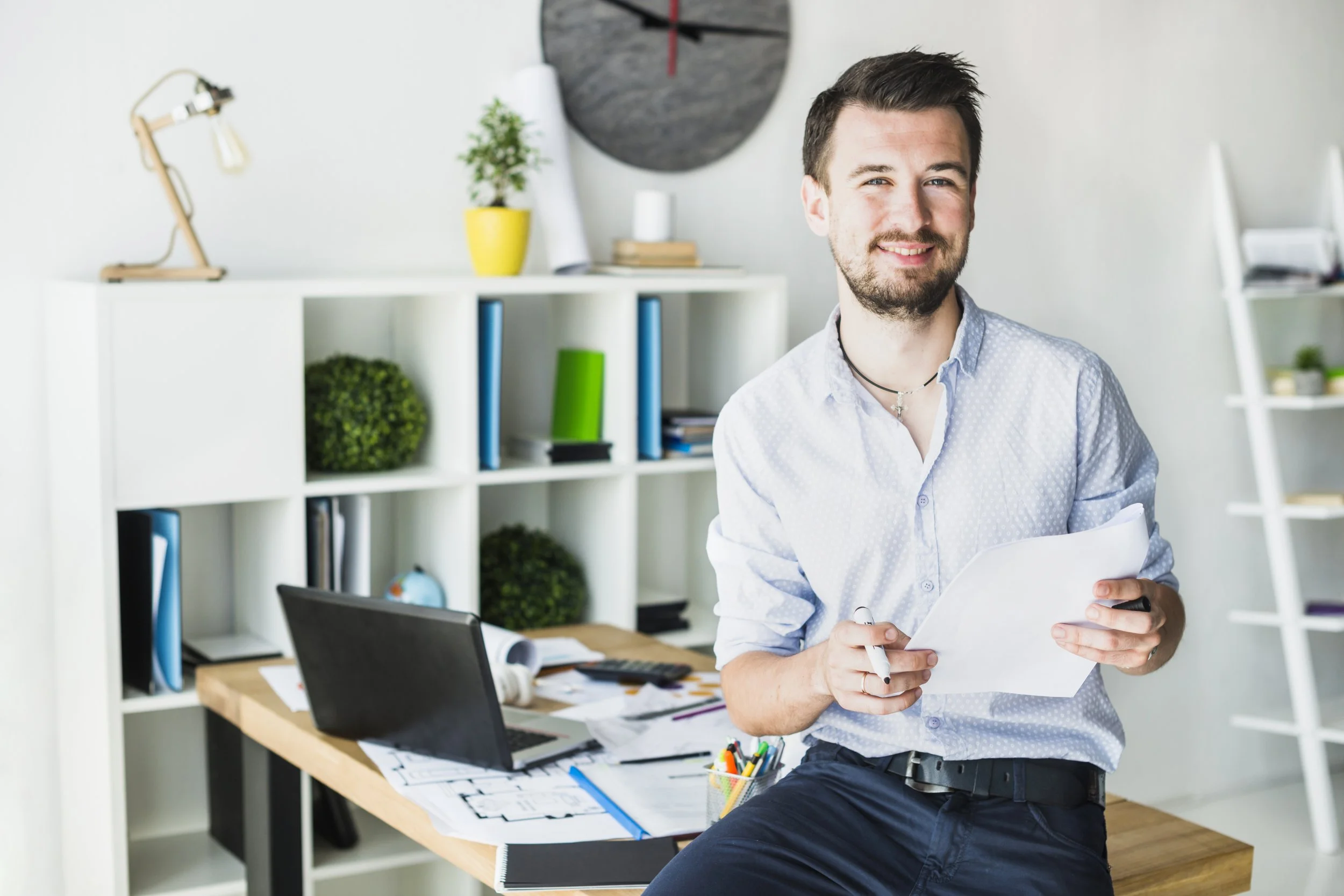 Smiling young man with a beard, holding papers and a pen in an office environment.