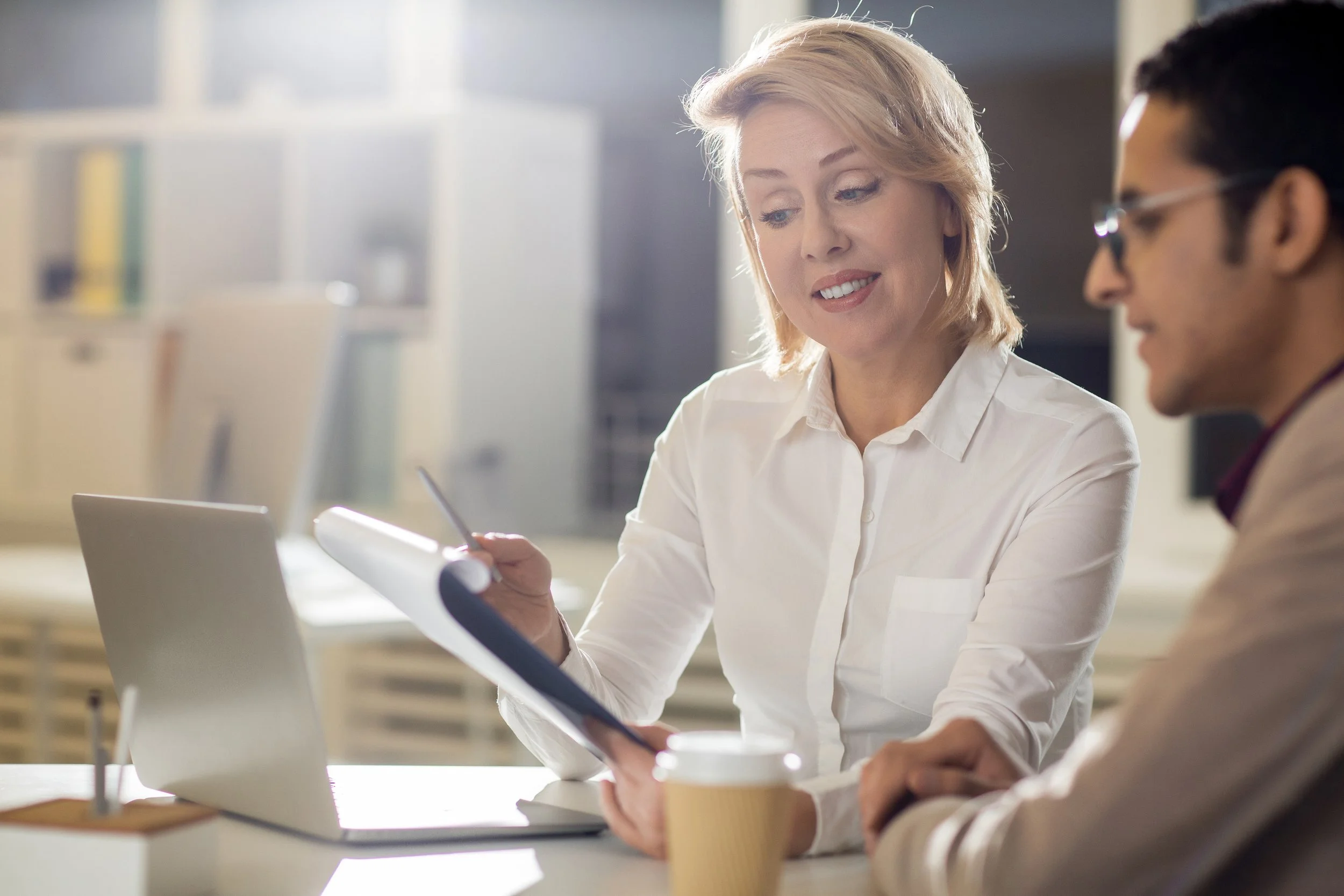 Two professionals, a woman with blonde hair and a man with dark hair and glasses, working together at a desk in an office. The woman is holding a pen and looking at documents, while the man has a disposable coffee cup in front of him. There is a laptop and other office supplies on the desk.
