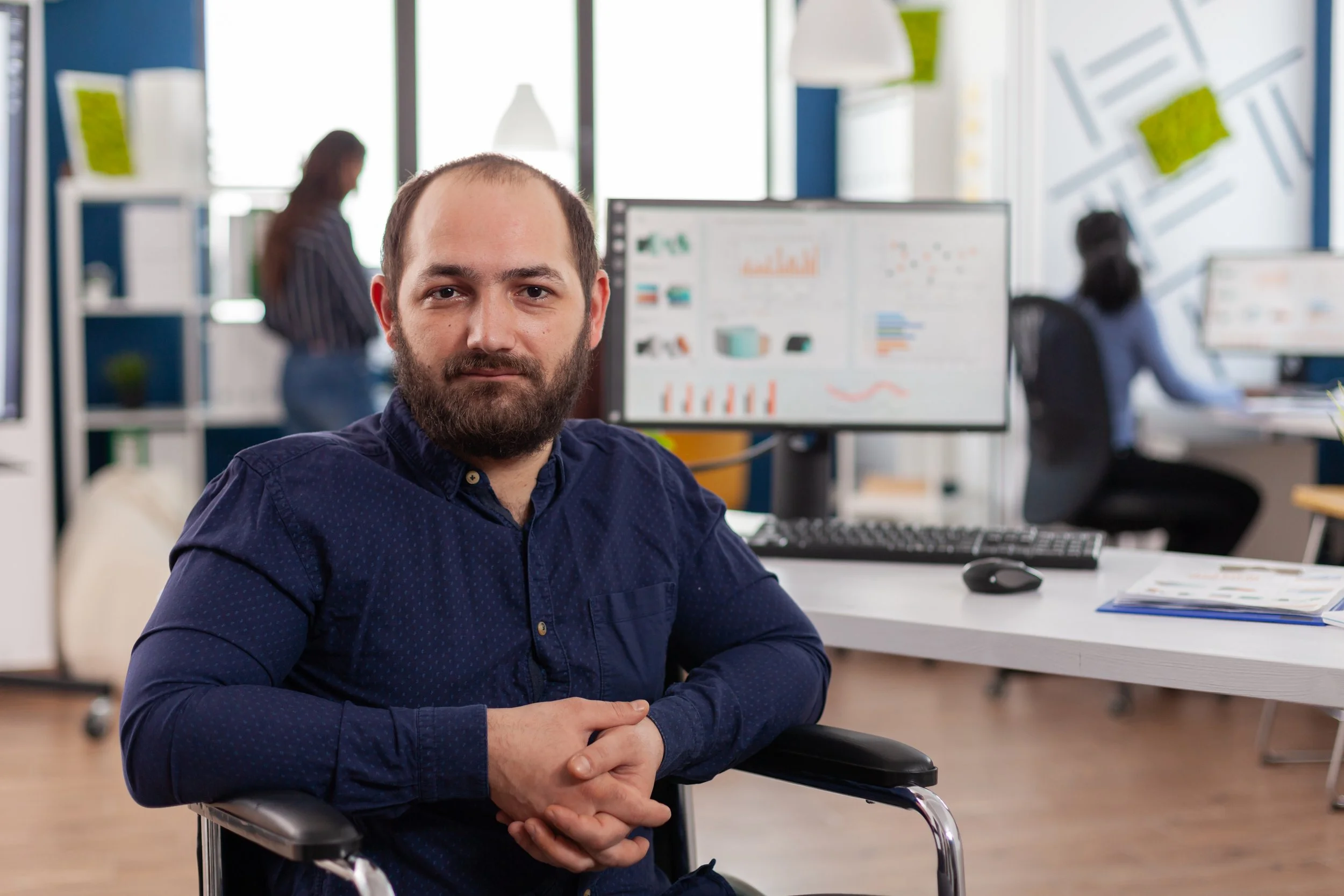 A man with a beard and short hair sitting in a wheelchair at a desk in an office. In the background, two women are working at desks with computer monitors, and there are large windows letting in natural light.