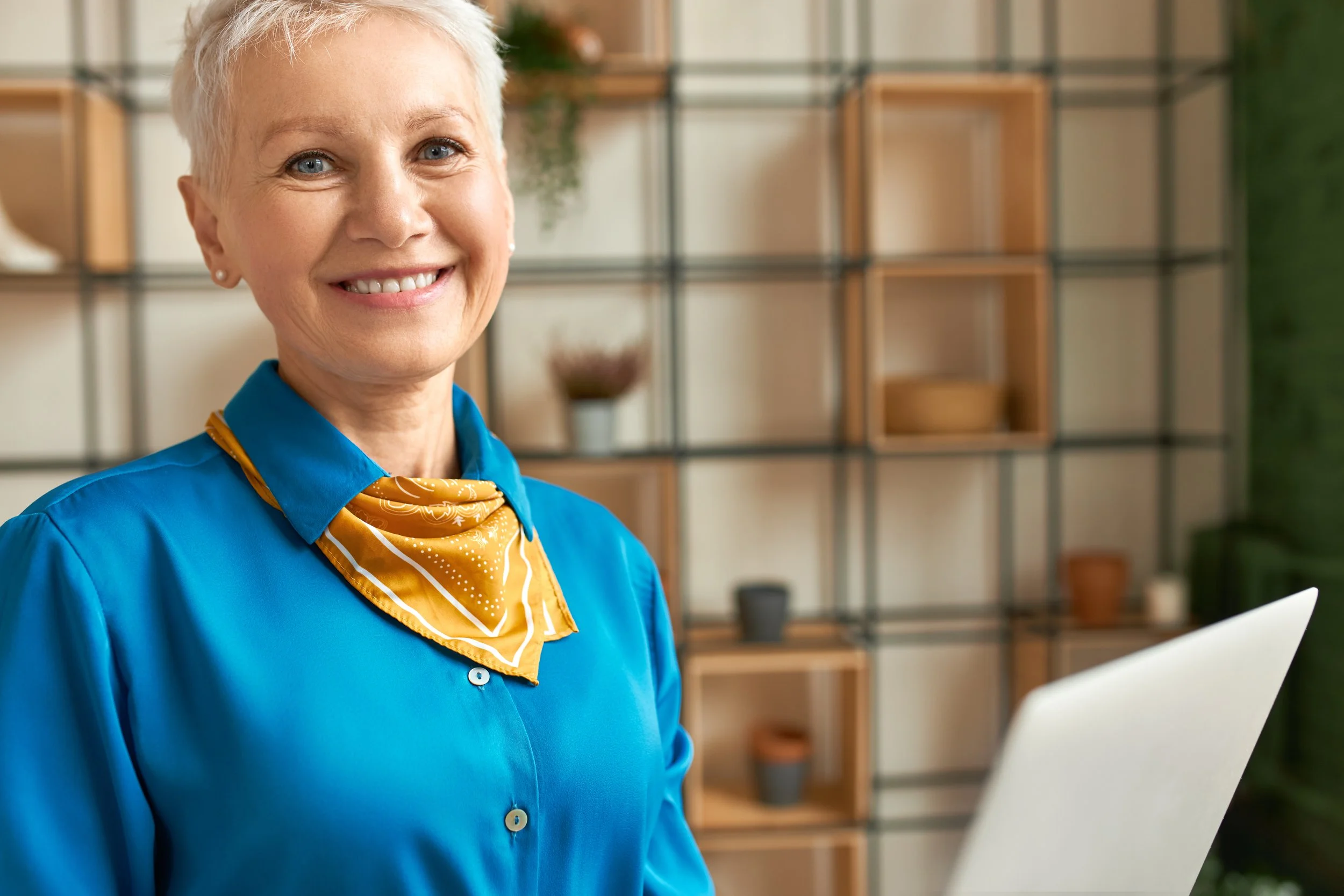 Smiling older woman with short gray hair, wearing a blue shirt and a yellow scarf, standing indoors in front of a shelving unit with potted plants and decorative items, holding a laptop.