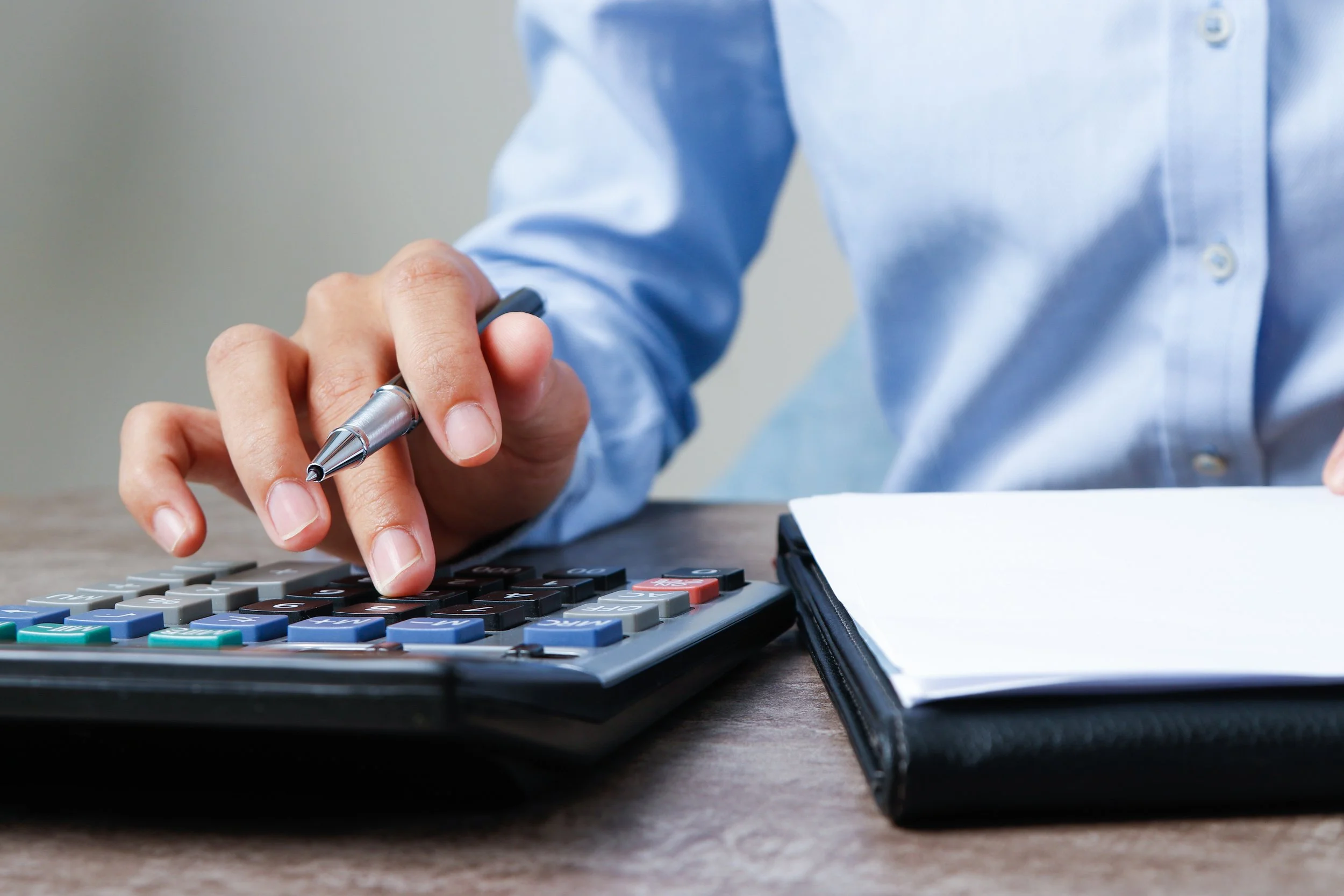 A person in a light blue button-up shirt is pressing the keys on a calculator with their right hand, holding a pen in the left hand, on a wooden desk with papers and a black folder.