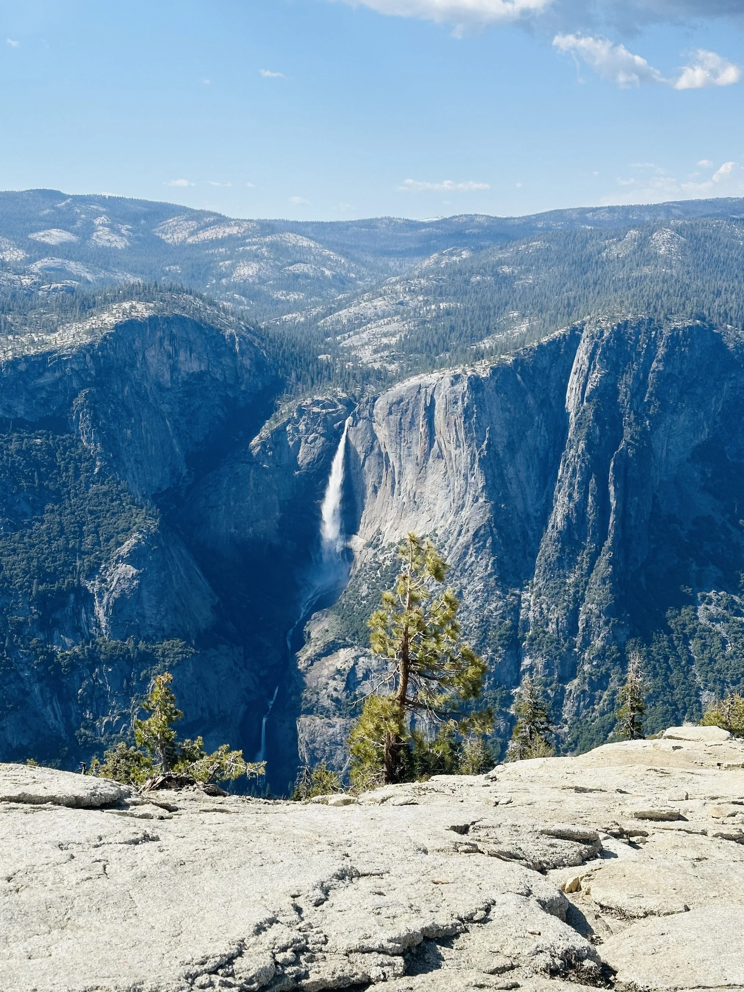 A scenic view of a waterfall cascading down a rocky cliff into a deep canyon, surrounded by pine trees and distant mountains under a blue sky with a few clouds.