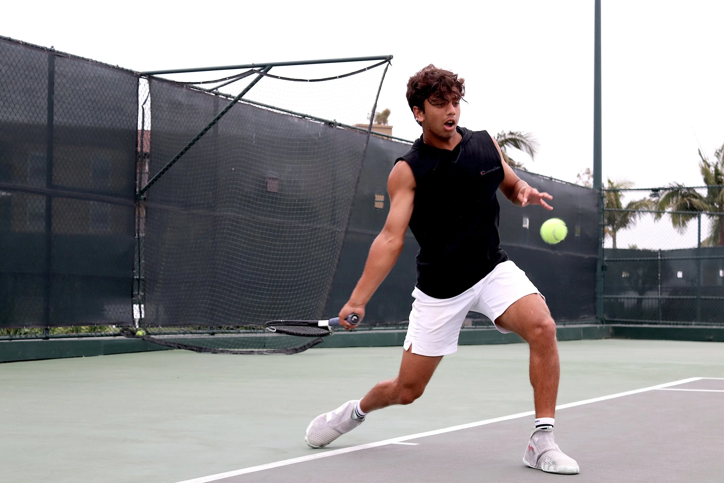 A young man playing tennis on an outdoor court, wearing a black sleeveless shirt, white shorts, and white sneakers, about to hit a tennis ball with a racket.