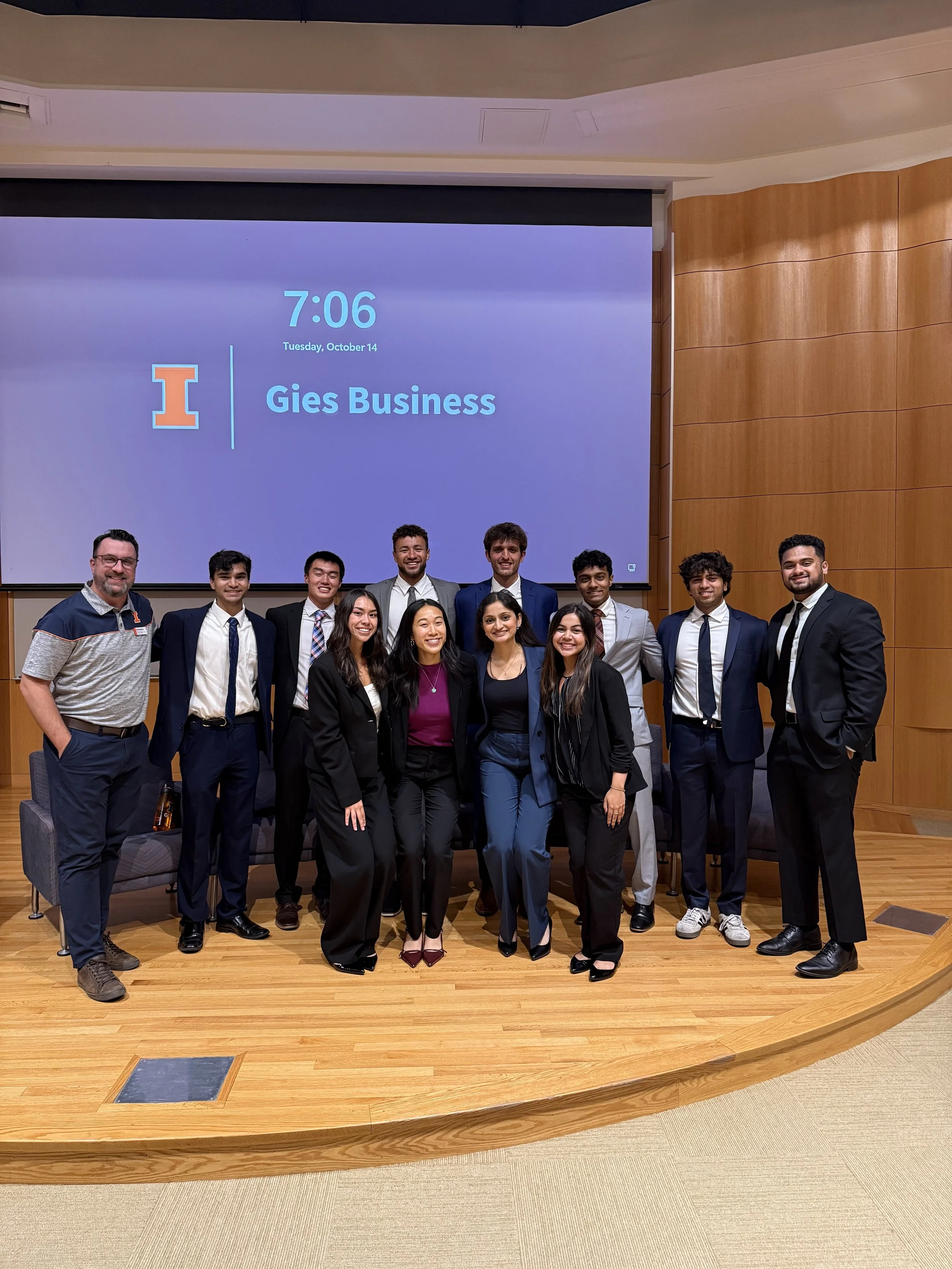 Group of students and a professor posing for a photo on a wooden stage in a conference room. The background features a large screen displaying a blue slide with the time 7:06, the date October 14, and a university logo with the text 'Gies Business'.